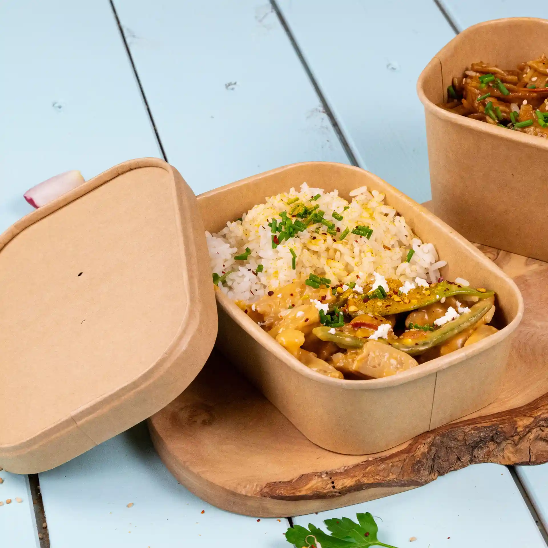 Rectangular kraft food container with rice and vegetables, partially open, on a wooden surface.