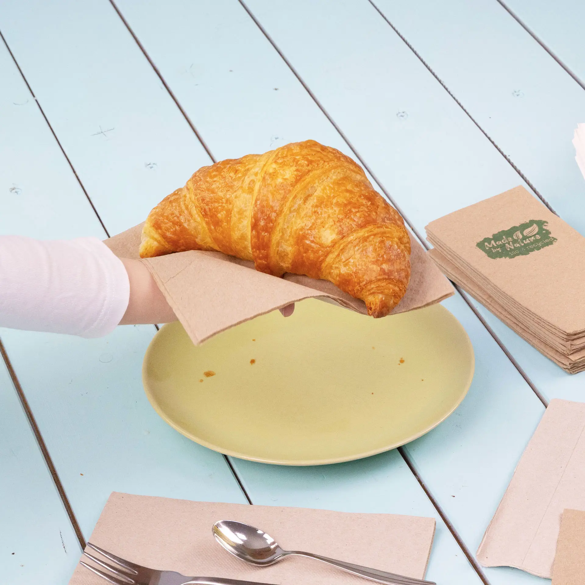 Eine Hand hält ein Croissant auf einer Papierserviette über einem gelben Teller auf einem blauen Tisch.