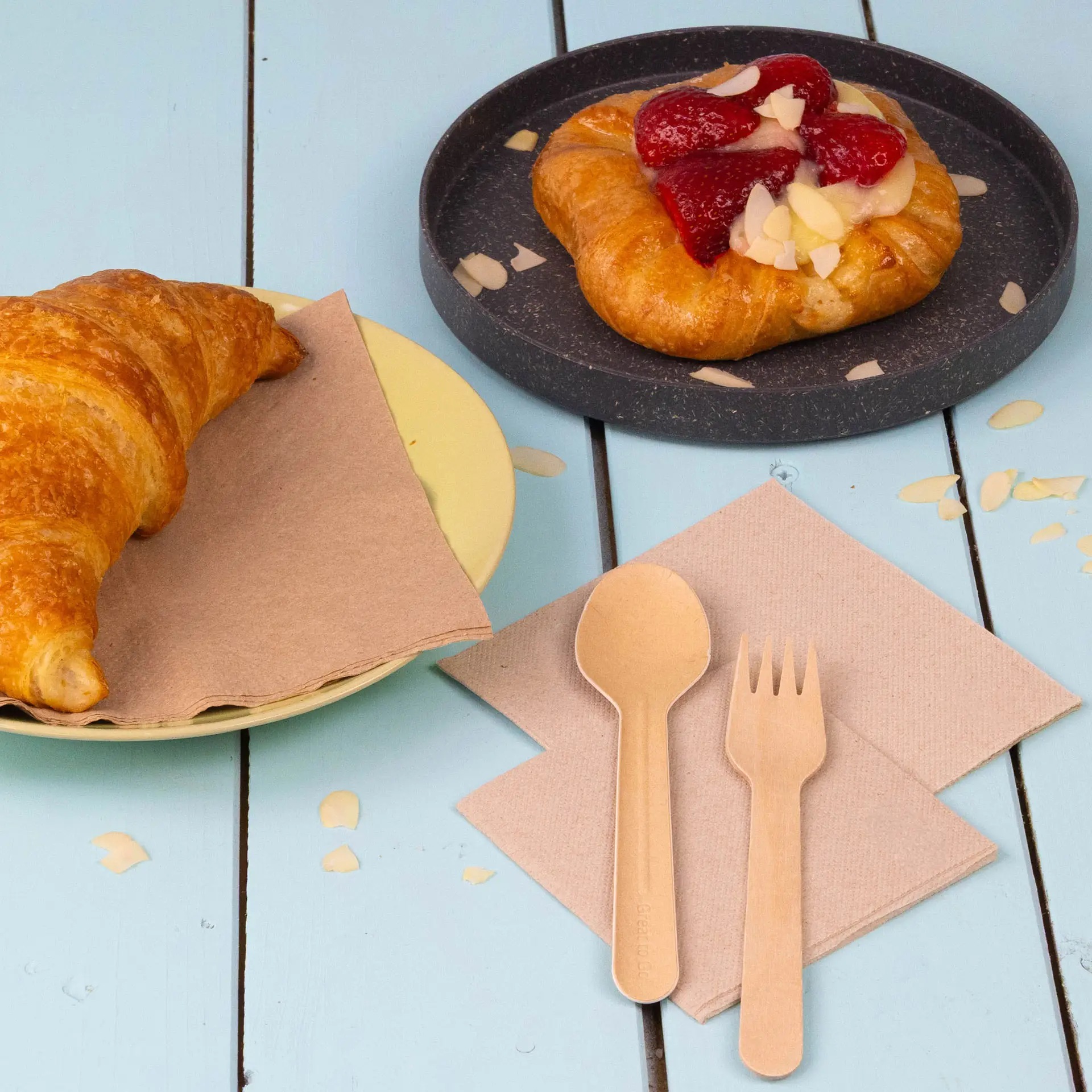 Image of a croissant on a yellow plate, a pastry with strawberries on a dark plate, and wooden cutlery on unbleached napkins.