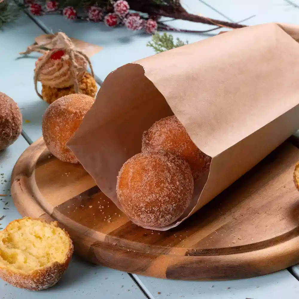 Eine braune Papiertüte enthält mehrere runde, mit Puderzucker bestäubte Donuts auf einem Holzschneidebrett, einen Donut teilweise geschnitten.