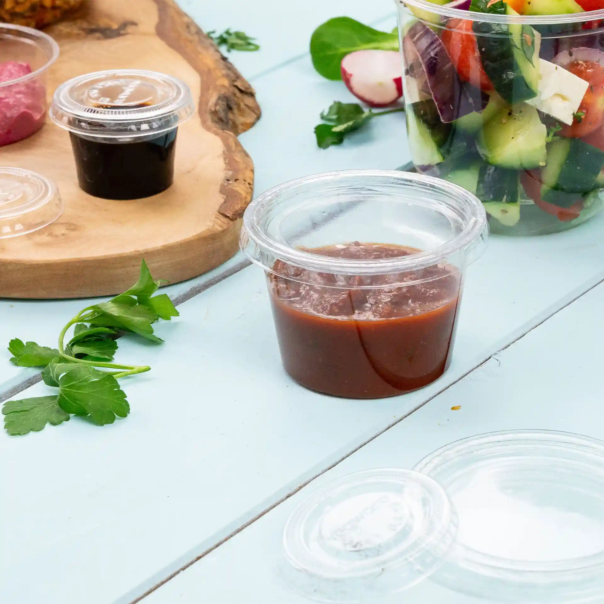 Clear PLA portion cup with a capacity of 80 ml, placed on a blue surface with parsley and other food items nearby.