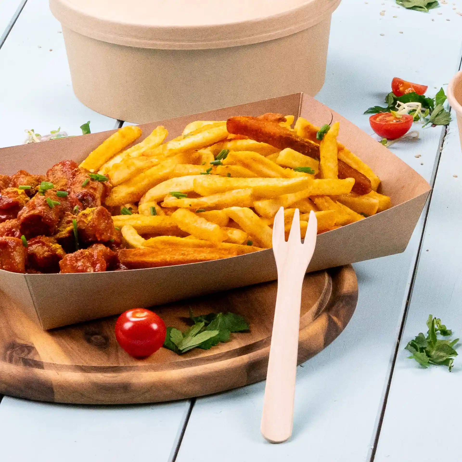 Image of a cardboard tray filled with fries and a meat dish, accompanied by a wooden fork and a cherry tomato on a wooden board.