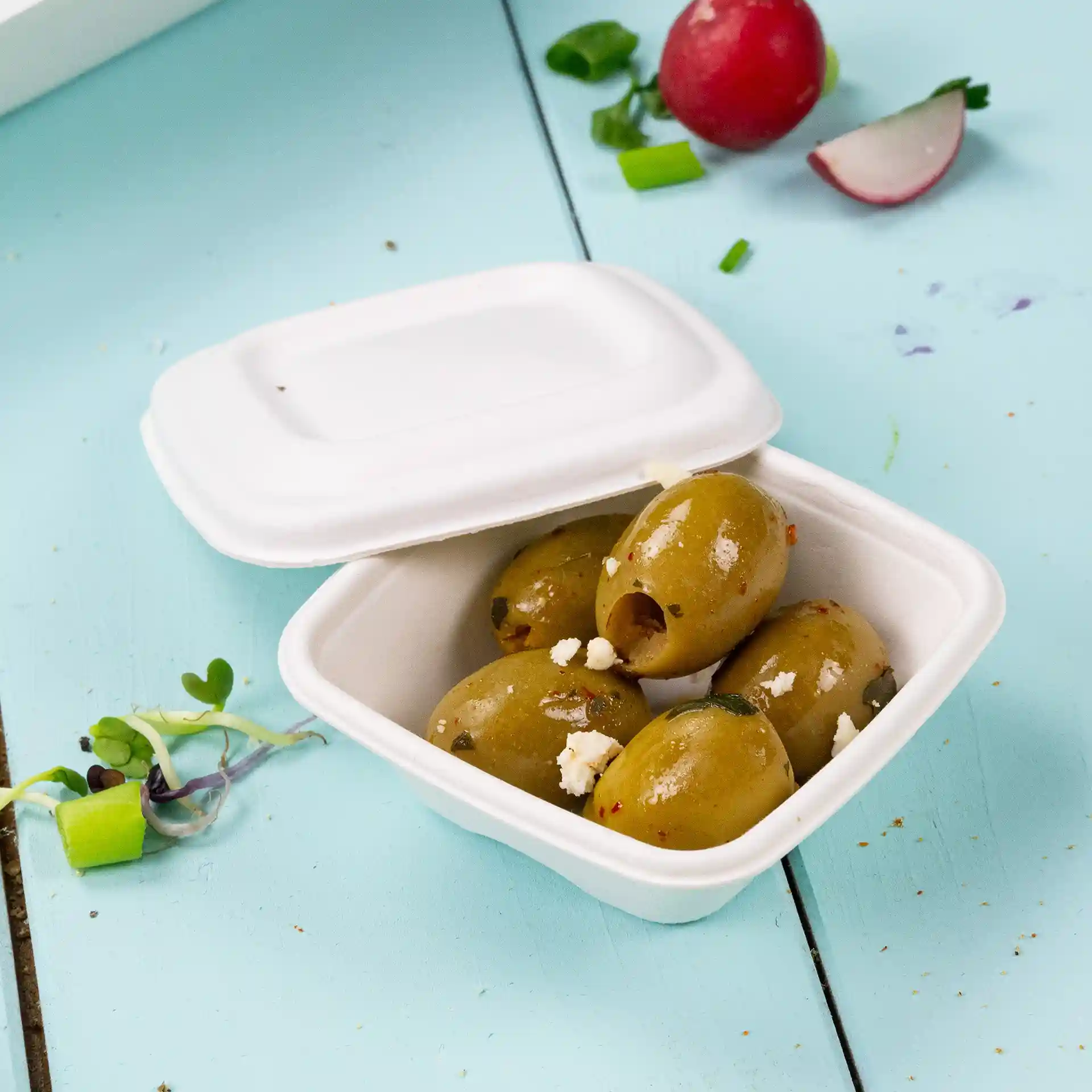 White square portion cup containing several green olives, placed on a light blue surface with scattered vegetables.