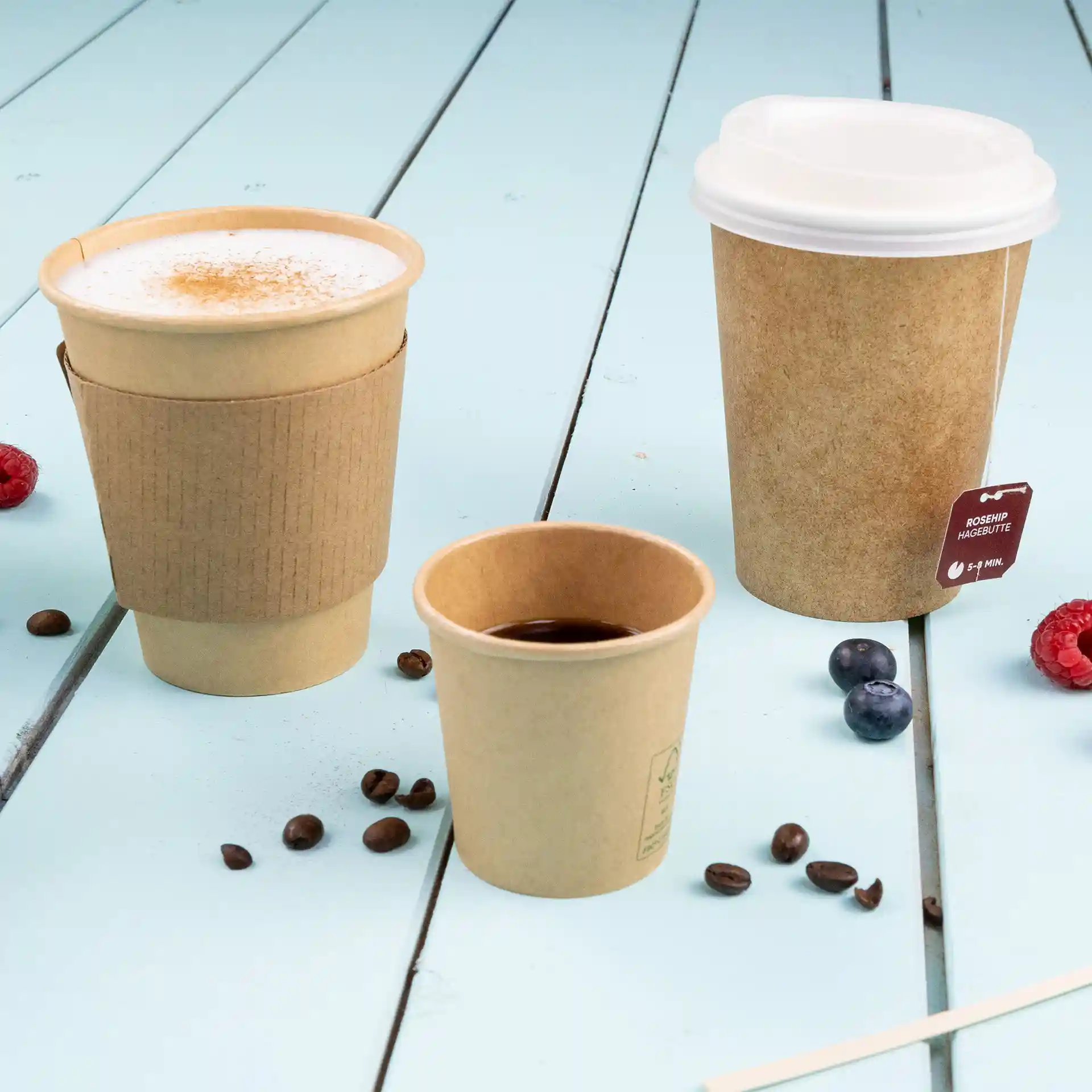 Bild von drei braunen Bechern in verschiedenen Größen auf einer blauen Holzoberfläche, mit Kaffeebohnen und Beeren verstreut.
