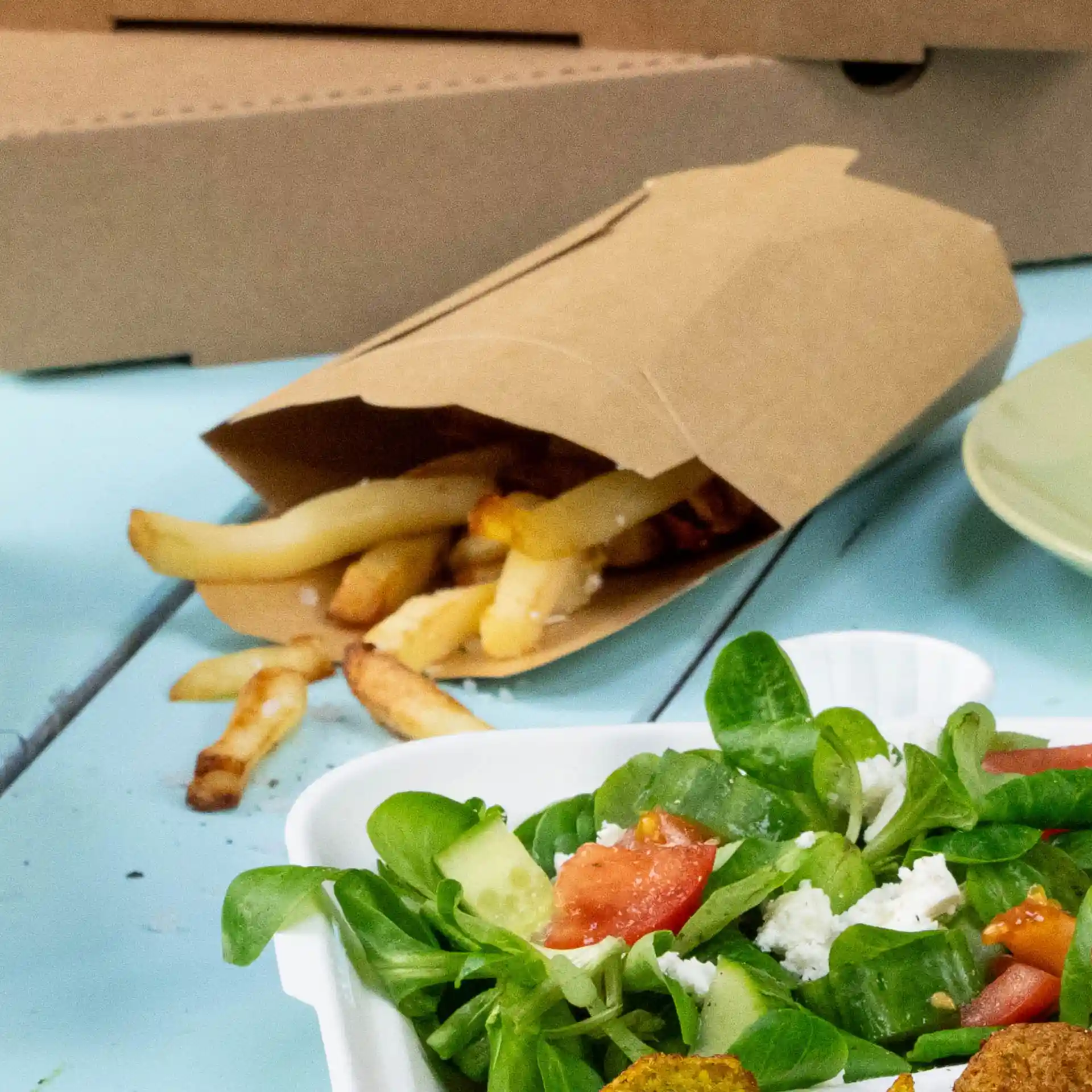 Brauner Karton-Chip-Scoop mit Pommes, daneben ein Salat in einem weißen Behälter auf einer blauen Oberfläche.