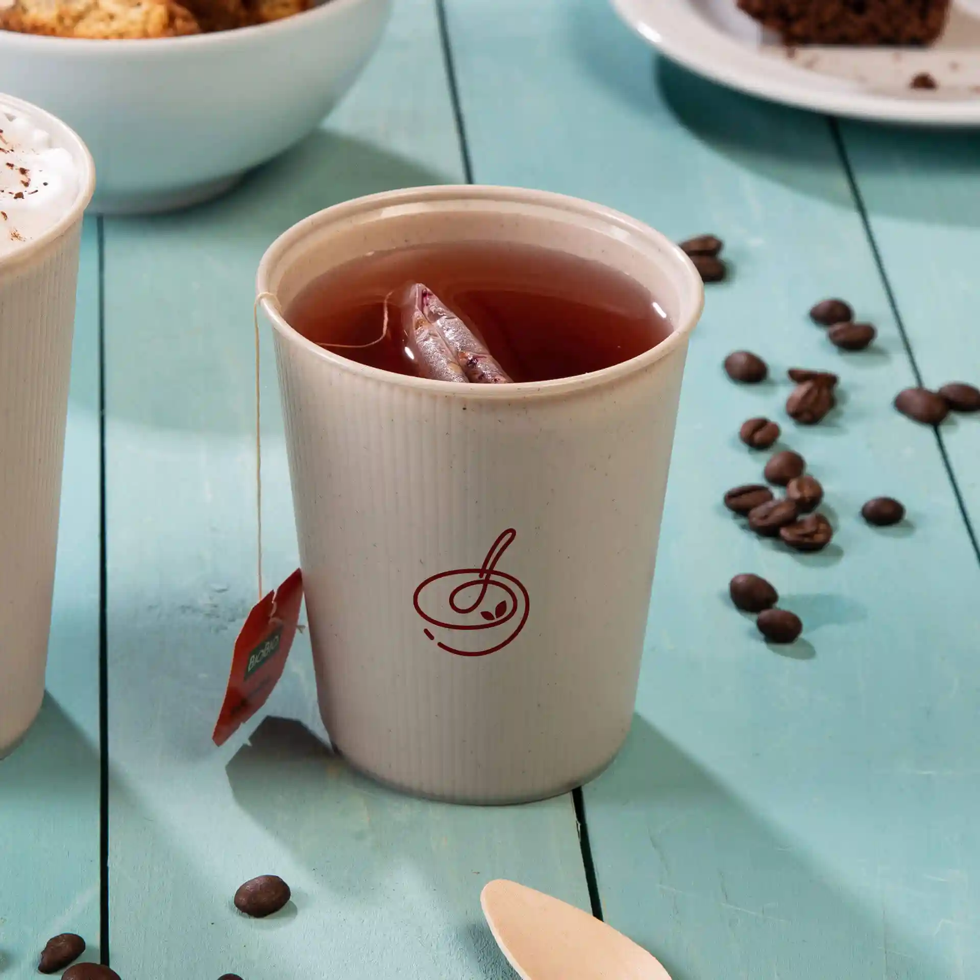 White reusable cup with a ripple design, filled with tea and a tea bag, on a blue wooden surface with coffee beans.