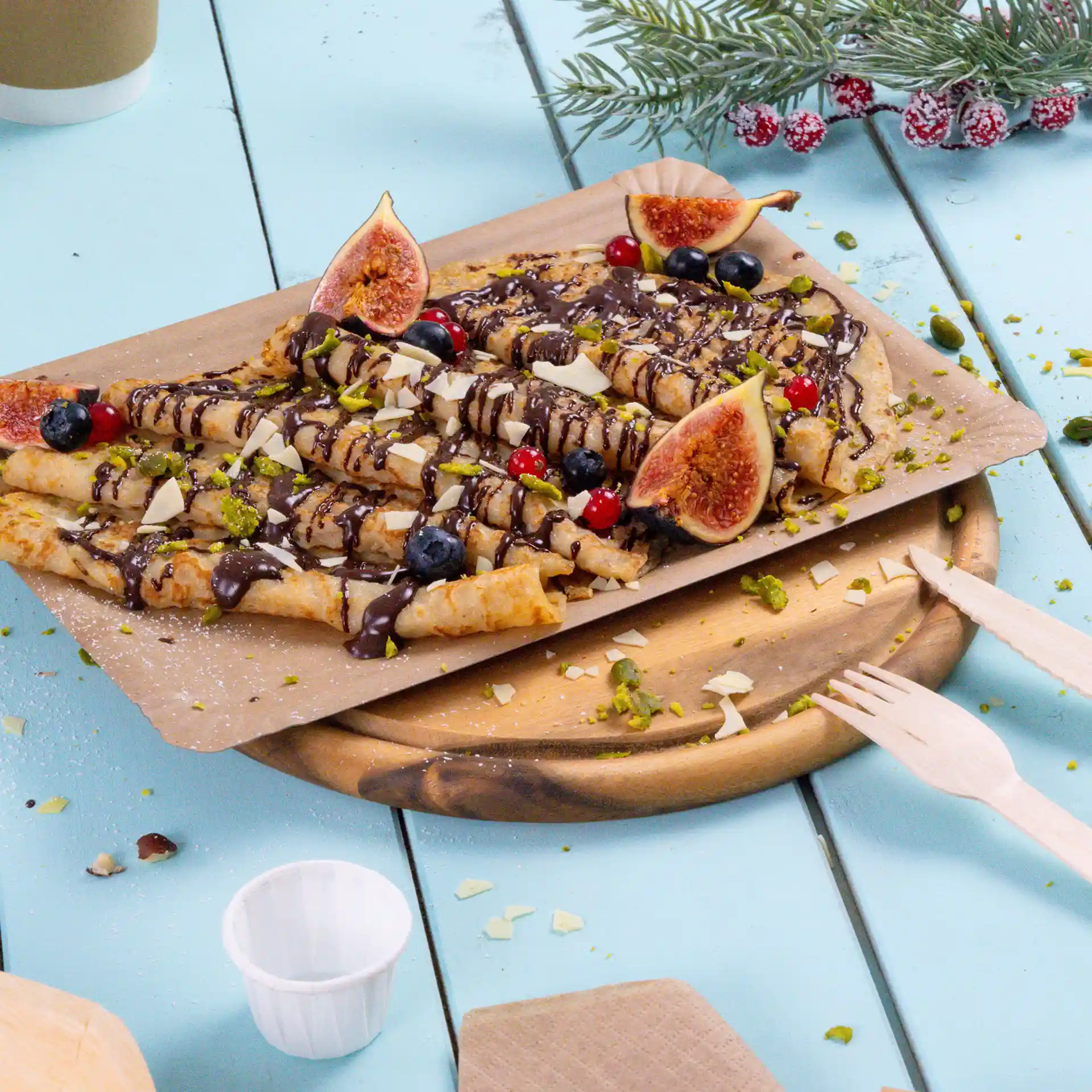 Rechteckiger brauner Pappteller mit gerollten Crêpes, belegt mit Schokolade, Feigen, Beeren und gehackten Nüssen.
