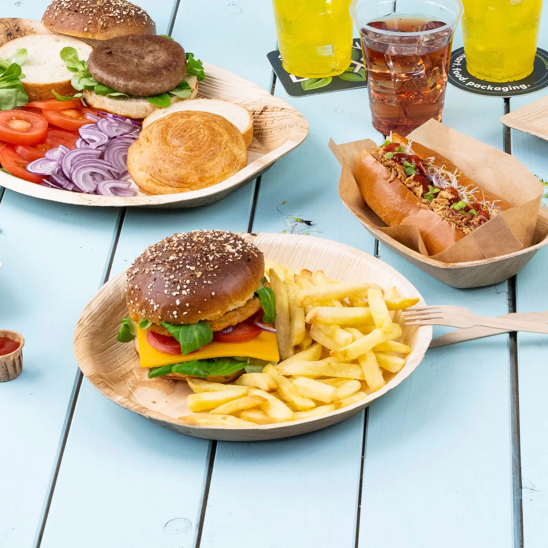A palm leaf plate holds a cheeseburger with lettuce, tomato, and fries, alongside slices of vegetables and a hot dog.