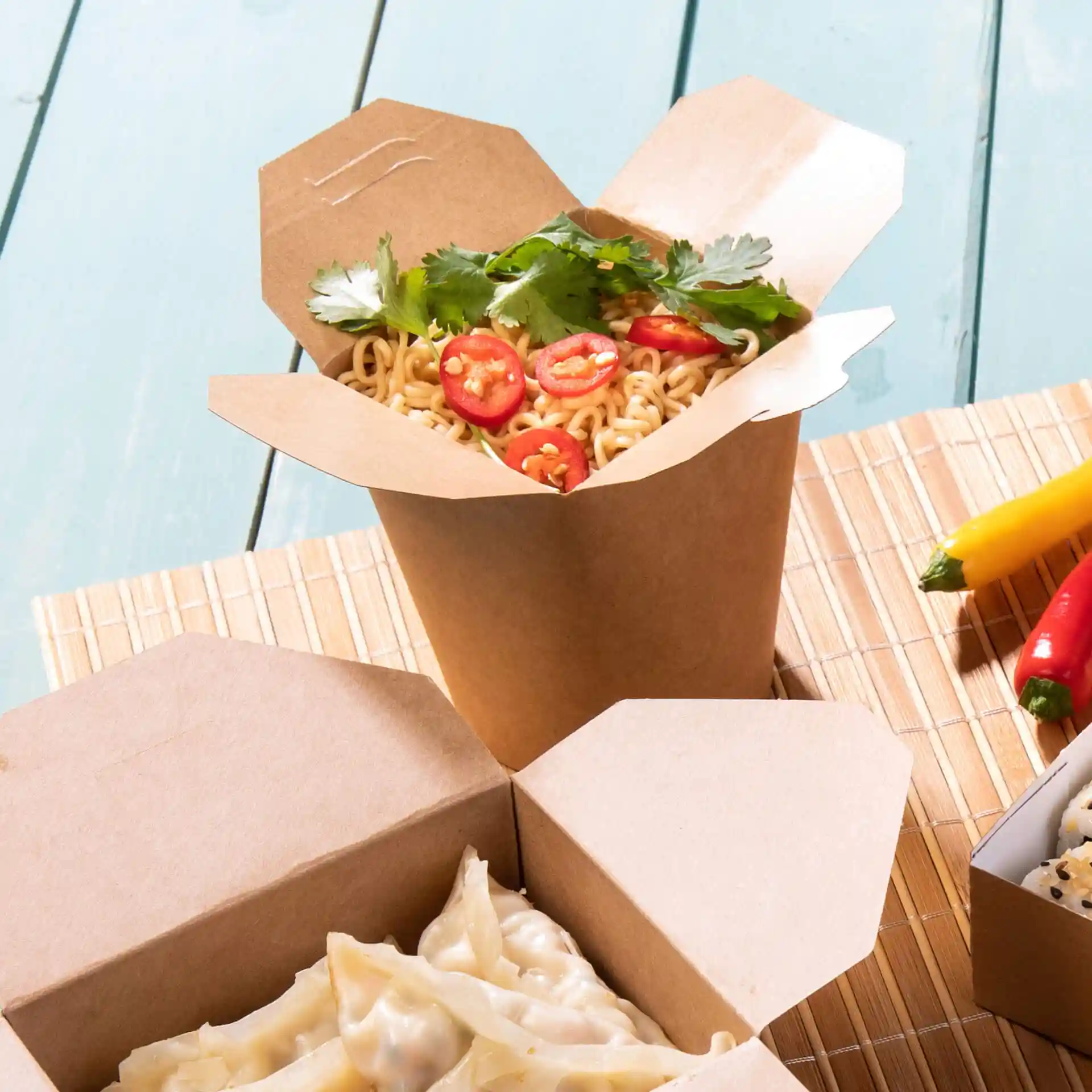 Brown cardboard noodle box filled with noodles, topped with sliced red chili and cilantro, on a bamboo mat.
