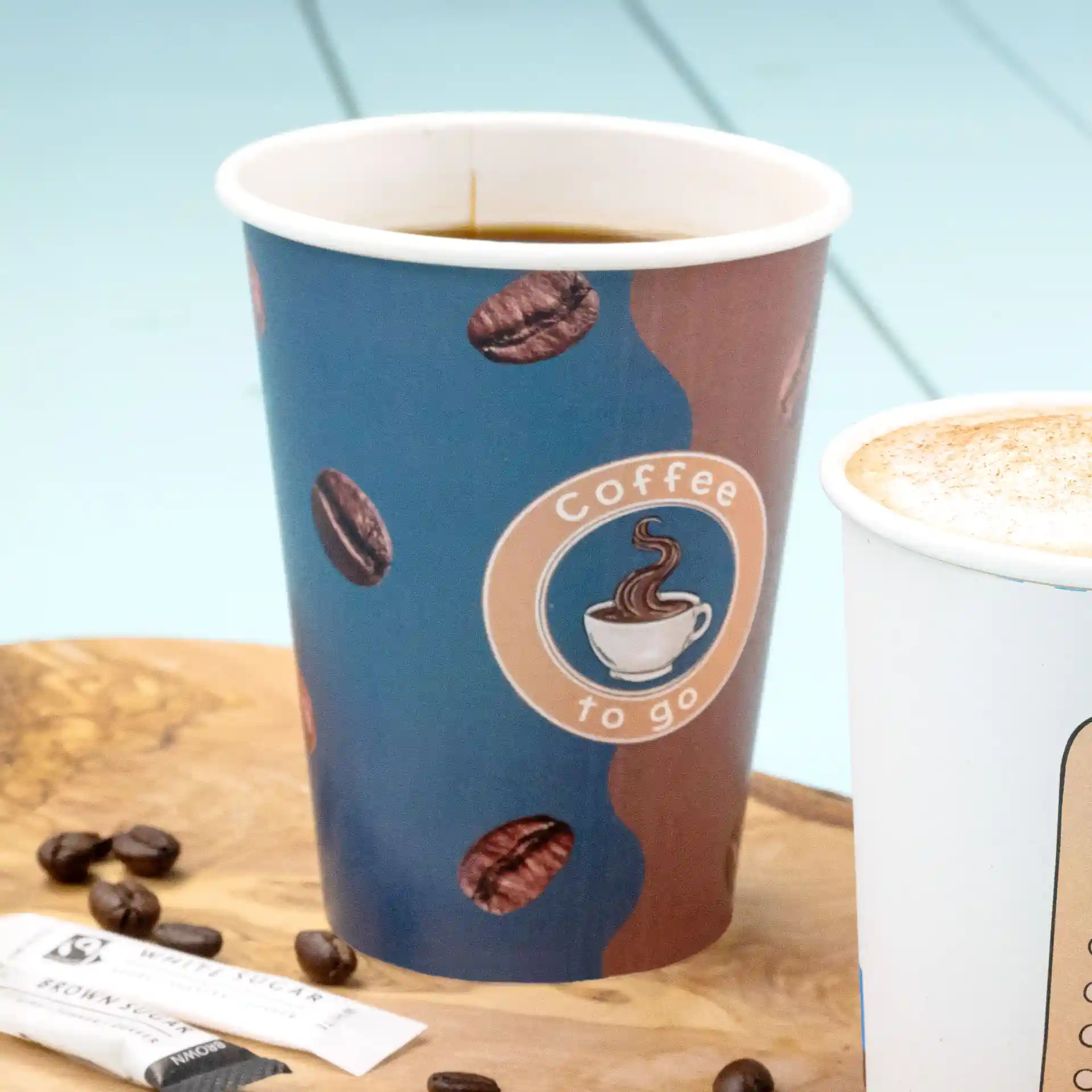 Paper coffee cup with printed design, filled with coffee, placed on a wooden surface with sugar packets and coffee beans.