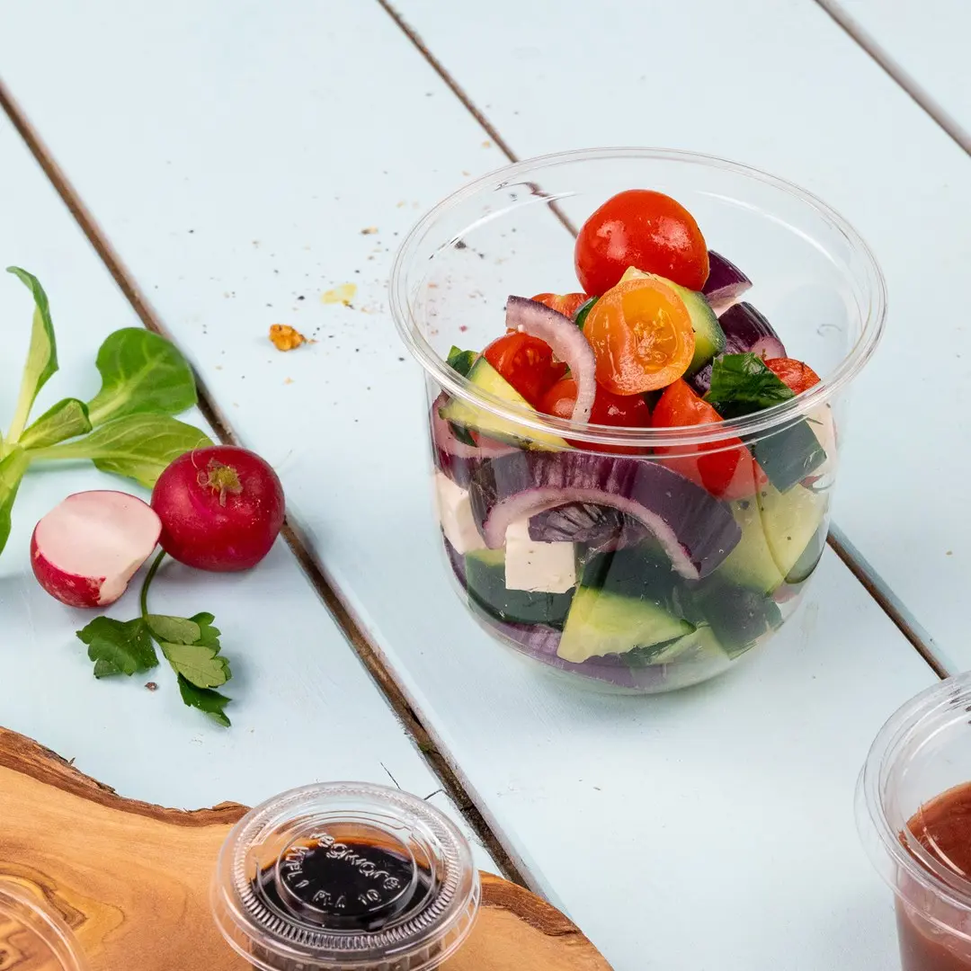 Klarer Plastikbecher gefüllt mit gemischtem Salat, einschließlich Kirschtomaten, Gurke, roter Zwiebel und Feta, mit Radieschen daneben.
