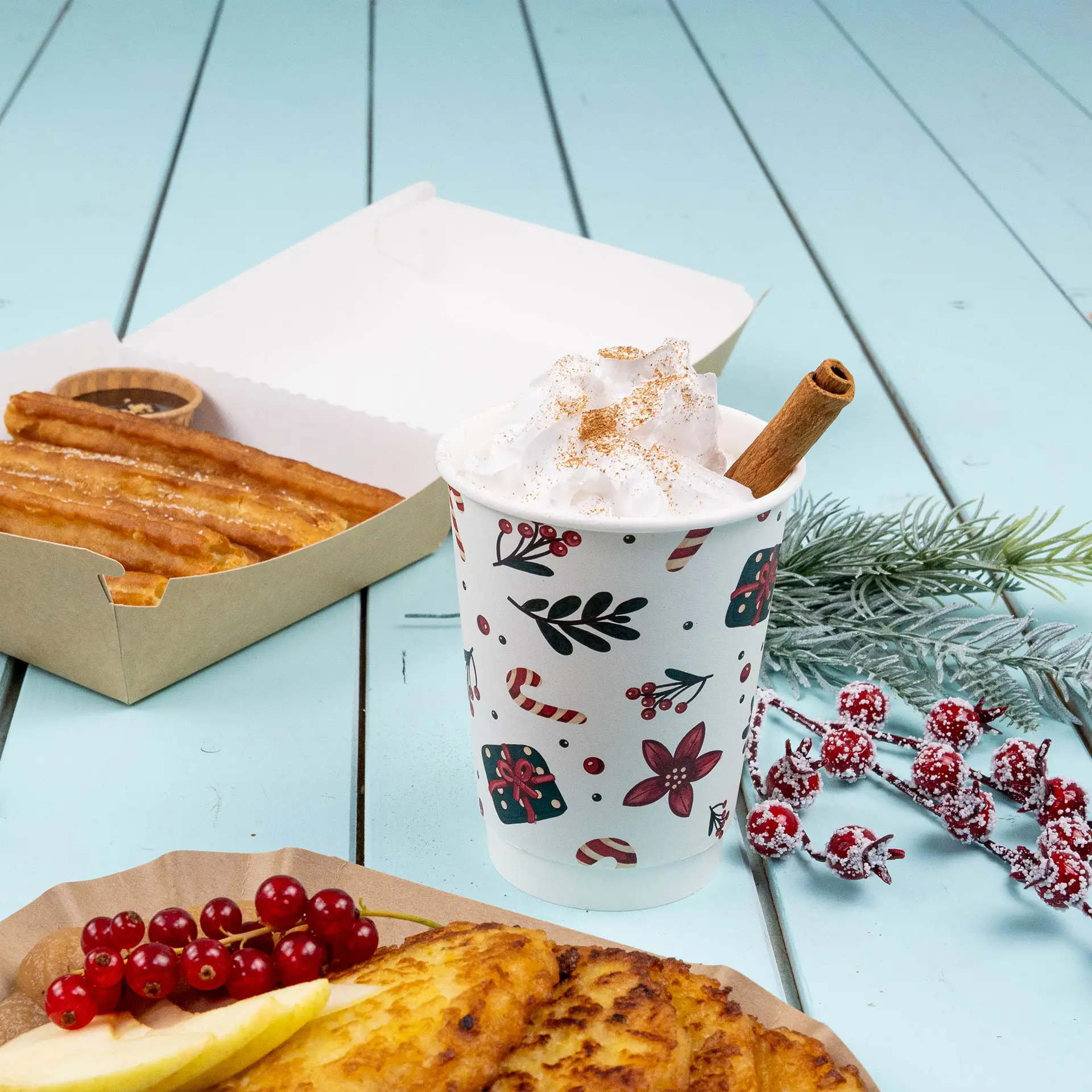 Double-wall coffee cup with winter design, filled with whipped cream and a cinnamon stick, alongside churros and fruit.