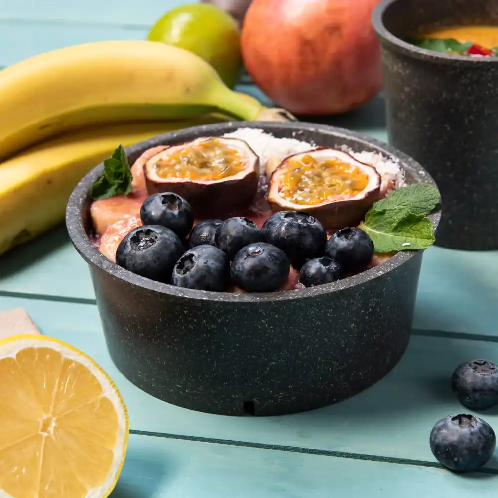 Eine schwarze Schüssel, gefüllt mit Früchten, darunter Heidelbeeren und Passionsfrucht, umgeben von Bananen und einer Zitronenscheibe.