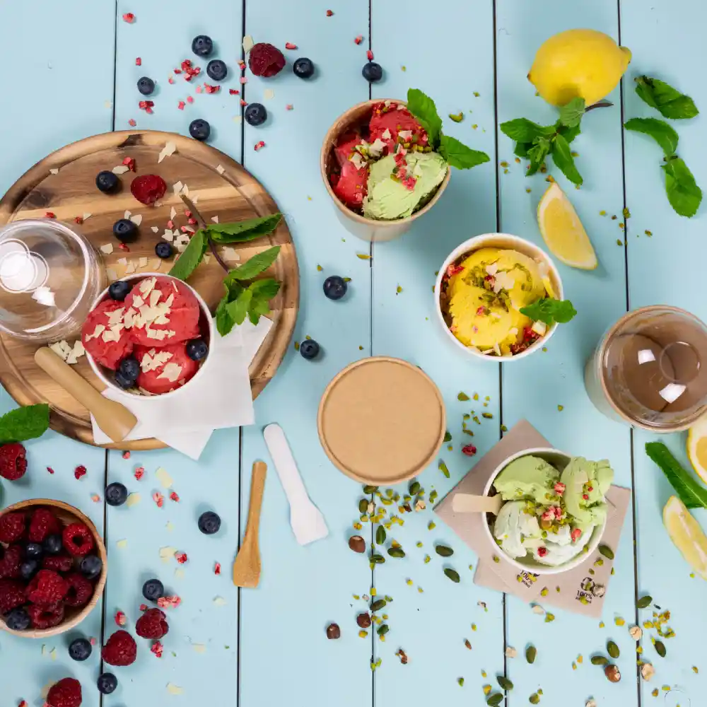 Colorful ice cream in cups with toppings on a blue wooden surface, surrounded by fresh fruits and utensils.