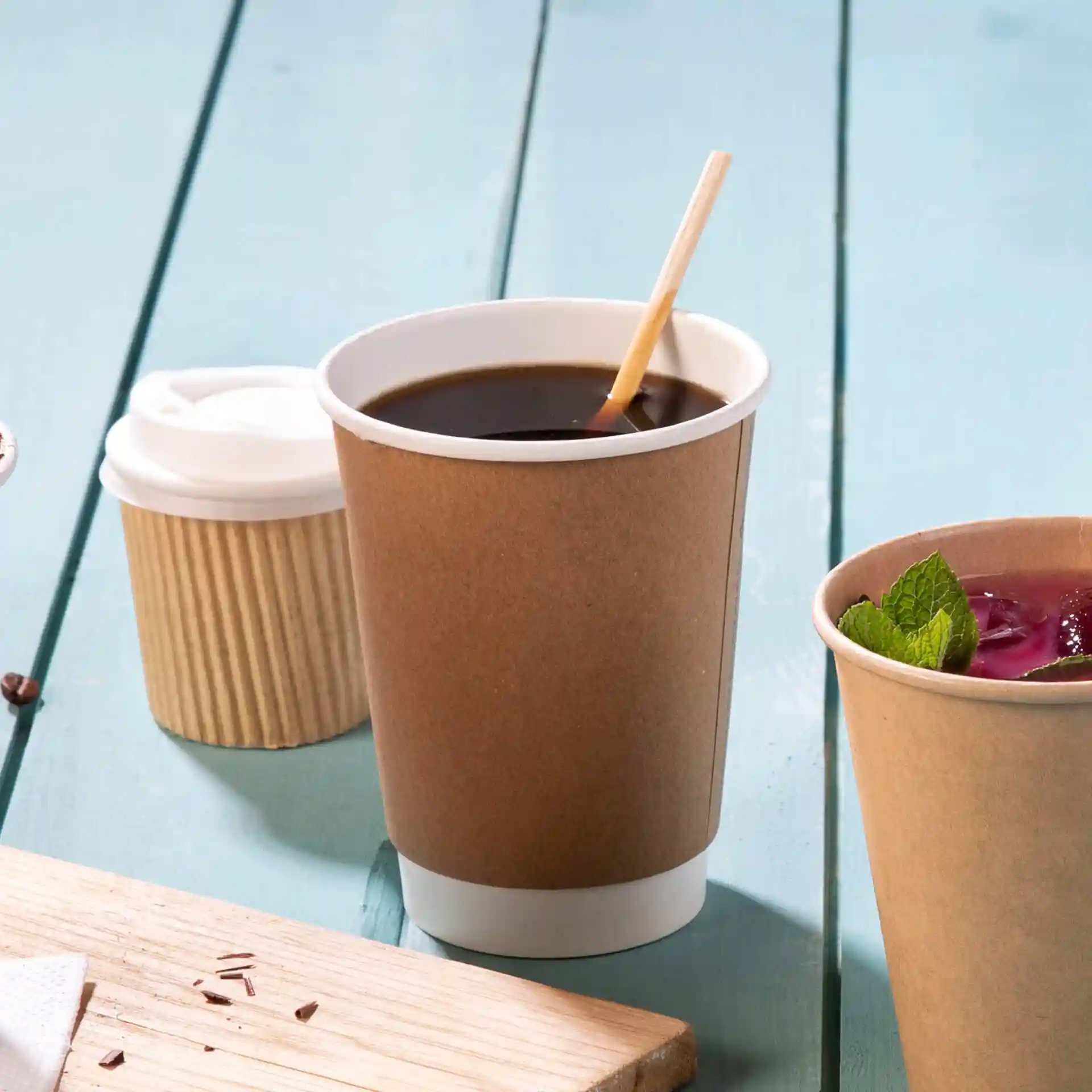 Brown paper cup with coffee and a bamboo stirrer, alongside a smaller cup and a wooden board on a blue surface.
