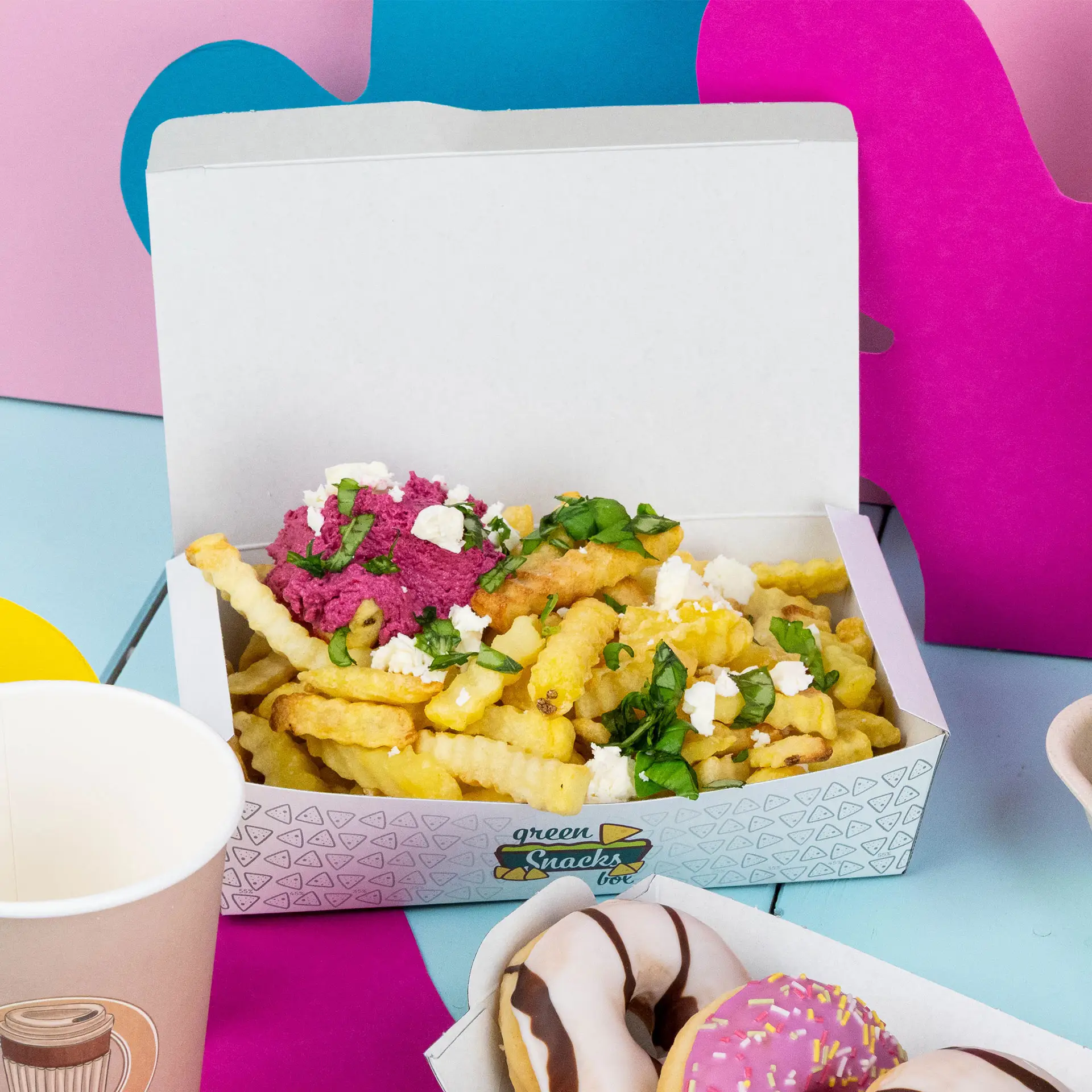 Eine weiße Snackbox gefüllt mit krummligen Pommes, bedeckt mit rosa Aufstrich, Feta und Kräutern, vor bunten Hintergründen.