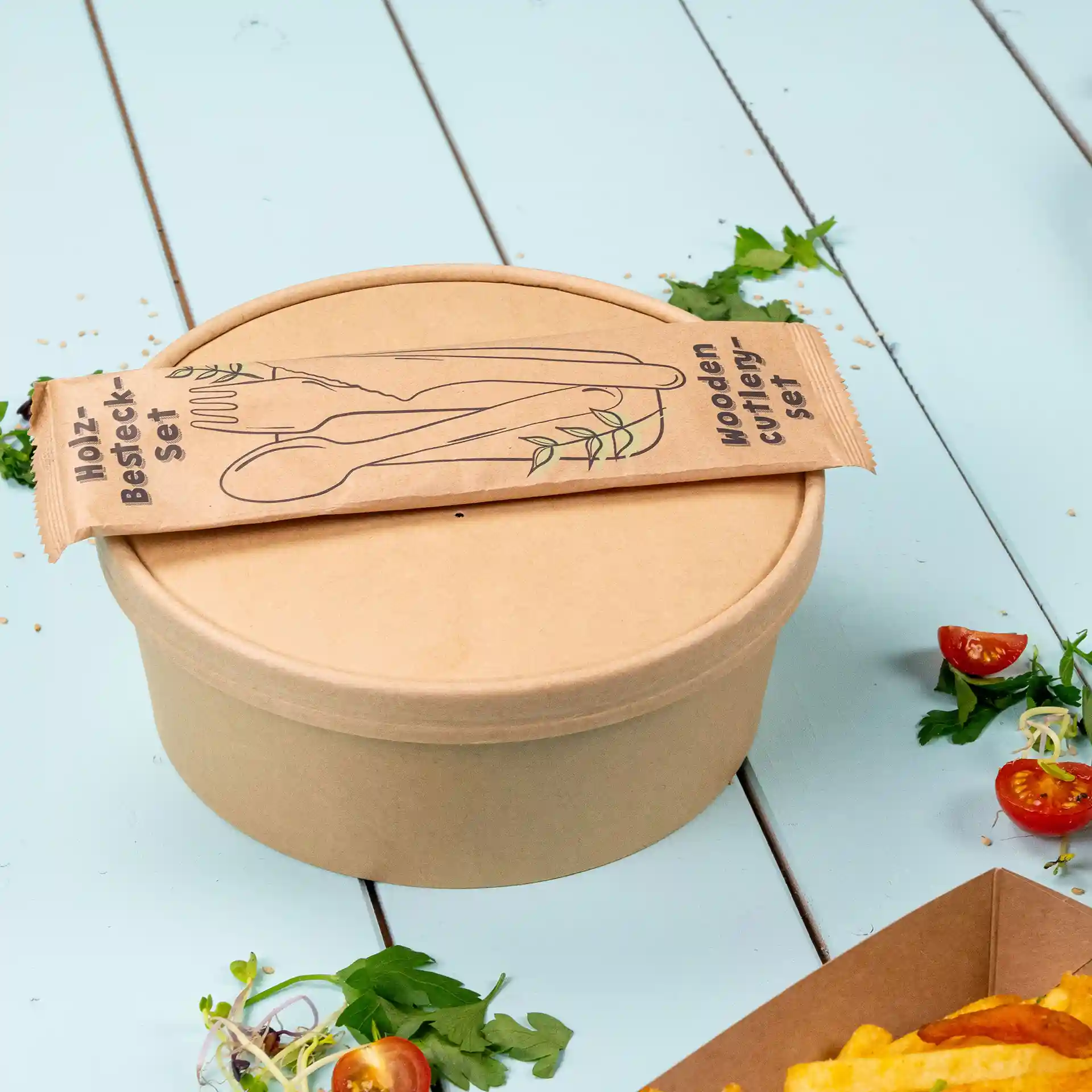 Brown round paper bowl with a wooden cutlery set wrapper on top, placed on a light blue surface.