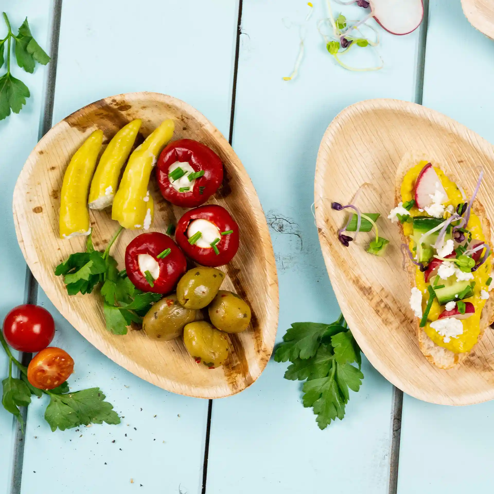 Two oval palm leaf plates with assorted vegetables, including yellow peppers, stuffed red peppers, and olives, on a blue background.