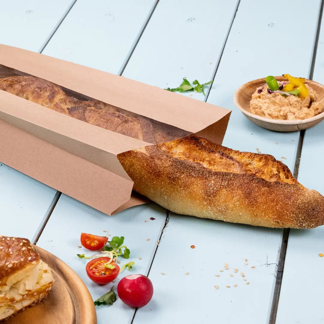 Brown paper bag partially open with a baguette, cherry tomatoes, radish, and a bowl of dip on a light blue surface.