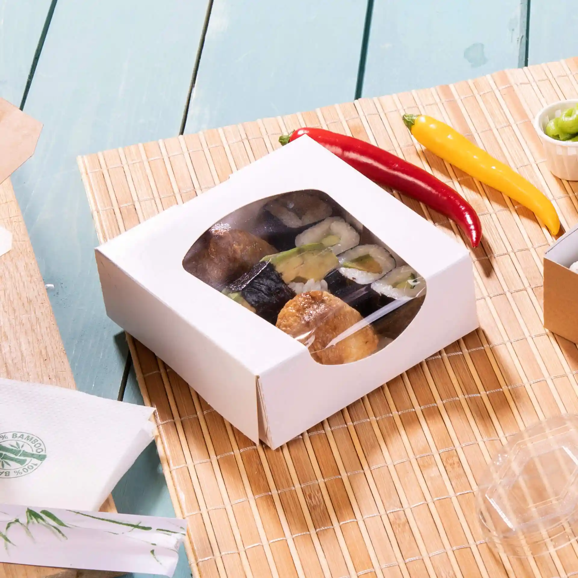 White cardboard window box containing sushi, placed on a bamboo mat with red and yellow chili peppers nearby.