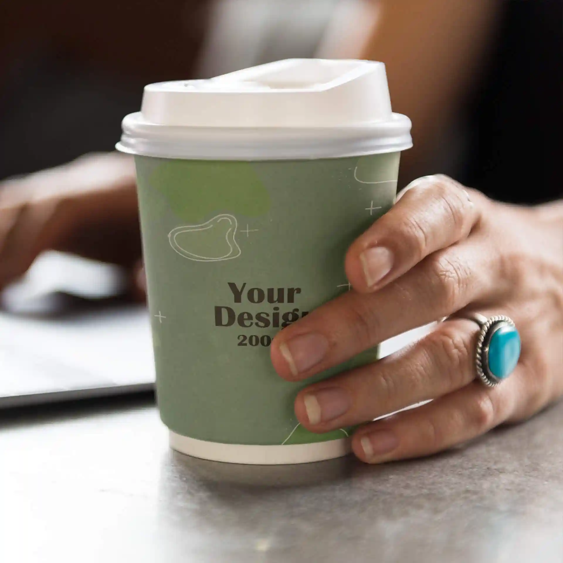 Hand holding a double-walled paper cup with a green design, featuring a white lid and a ring on the finger.