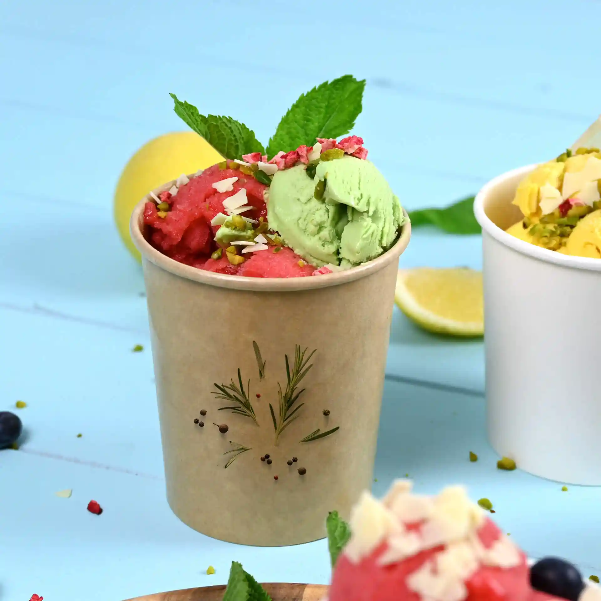 Brown cardboard ice cream cup filled with green and pink ice cream, topped with mint leaves and nuts.