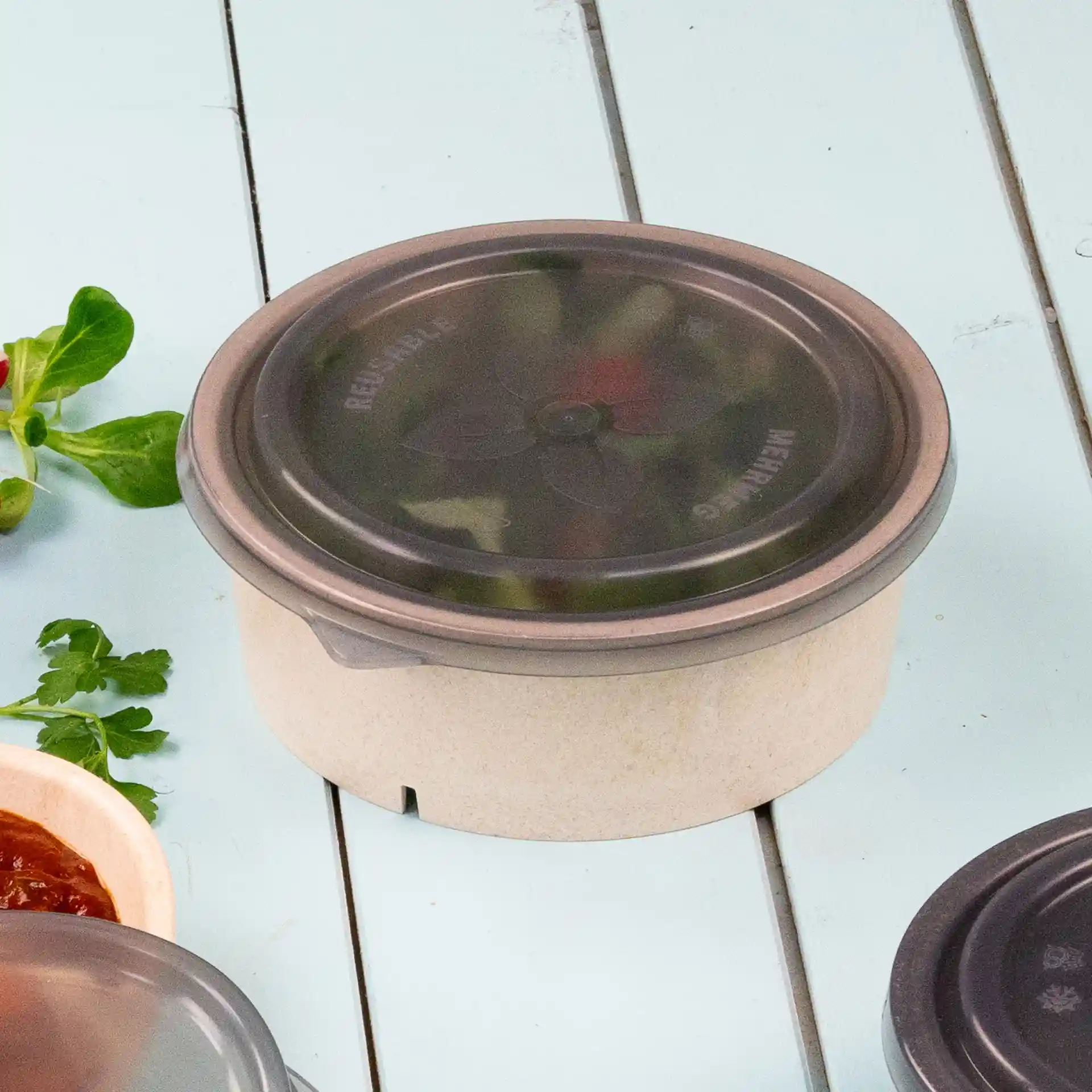 Round bowl with a grey reusable lid, placed on a light blue surface with herbs and sauce nearby.