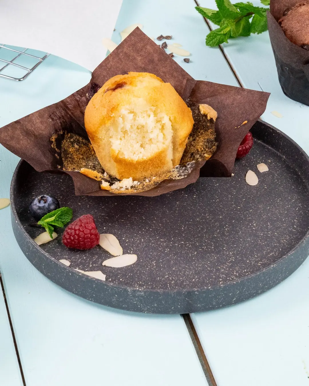 Muffin in a brown paper wrapper on a dark plate, surrounded by raspberries, blueberries, mint leaves, and almond slices.