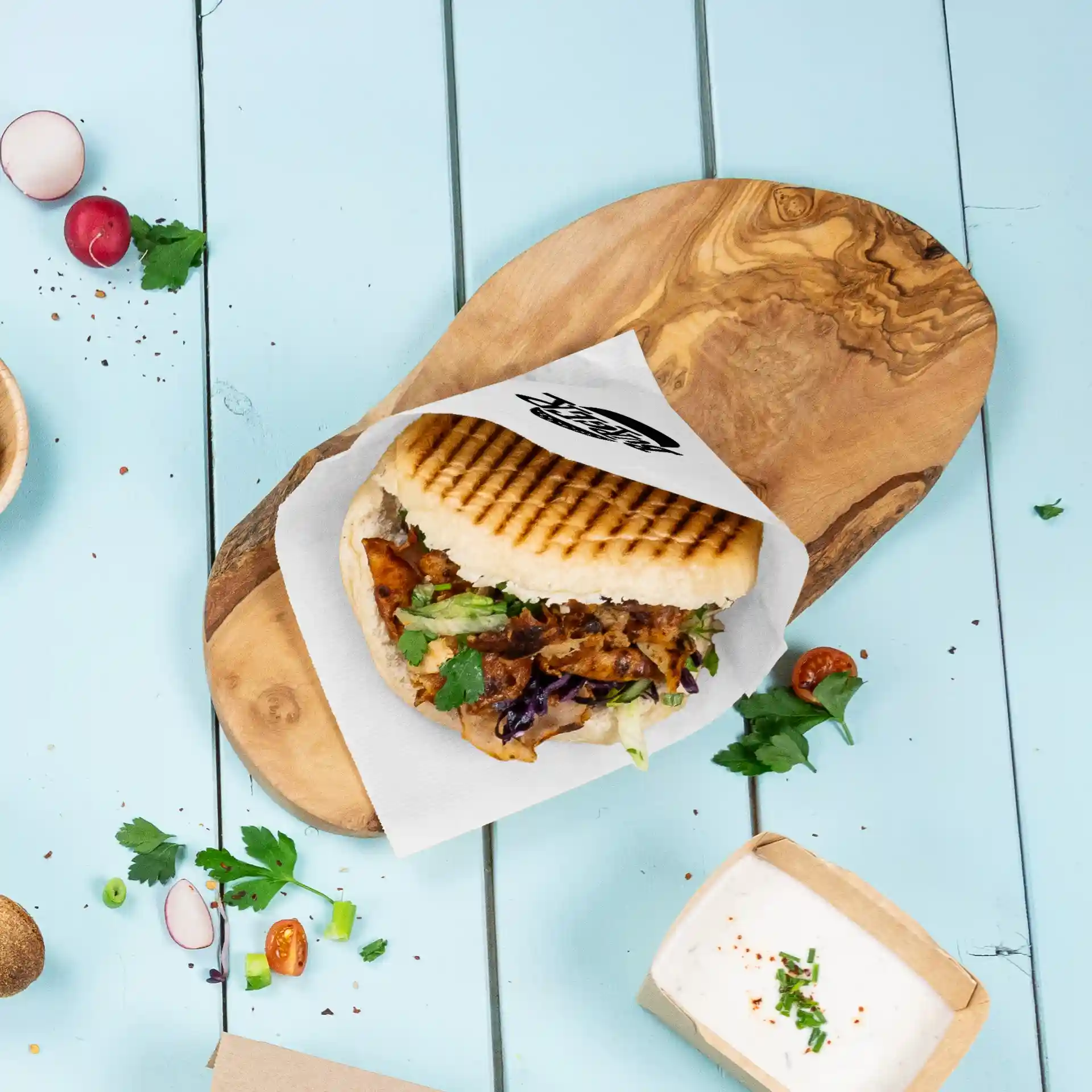 Bild eines Sandwichs auf einem Holzbrett mit einem bedruckten Sleeve, umgeben von frischem Gemüse und einem kleinen Behälter mit Sauce.