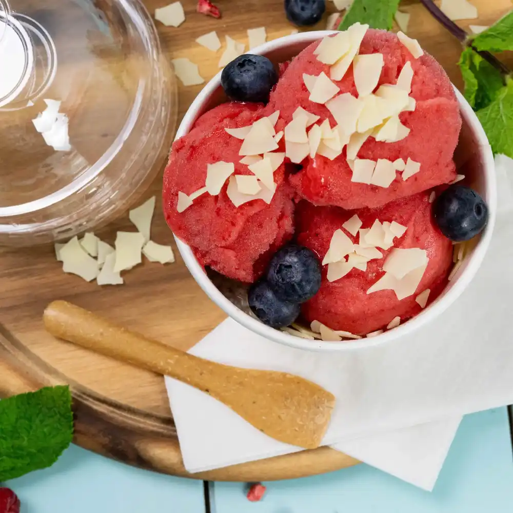 Bowl of red sorbet topped with chocolate shavings and blueberries, accompanied by a wooden spoon on a wooden tray.