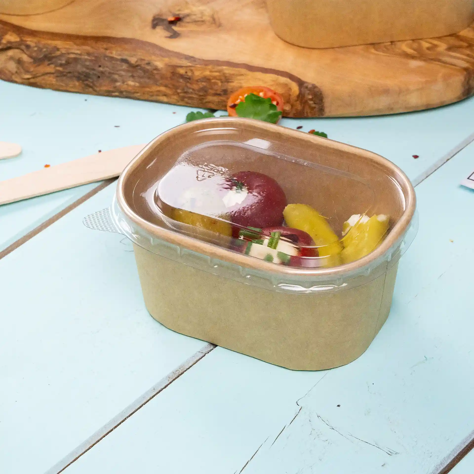 Rectangular kraft food container with a transparent lid, containing assorted vegetables, on a light blue surface.