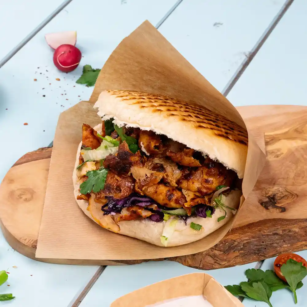 Ein gefülltes Pita-Sandwich mit gegrilltem Hähnchen, Salat, rotem Kohl und Koriander auf einem Holzbrett.