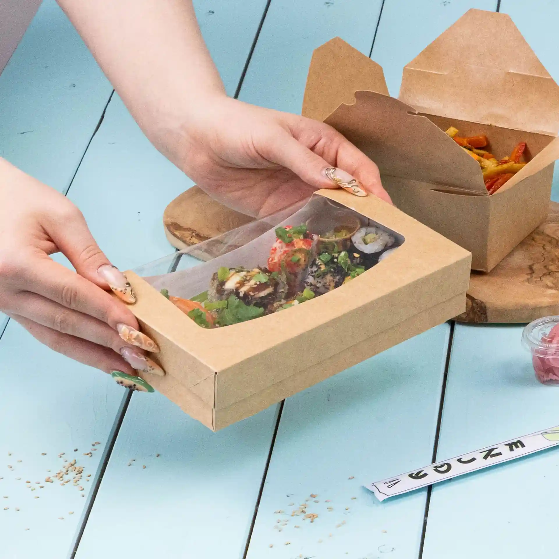 Kartonfensterbox mit transparentem Deckel, enthält Sushi und Salat, in Händen vor blauem Hintergrund.
