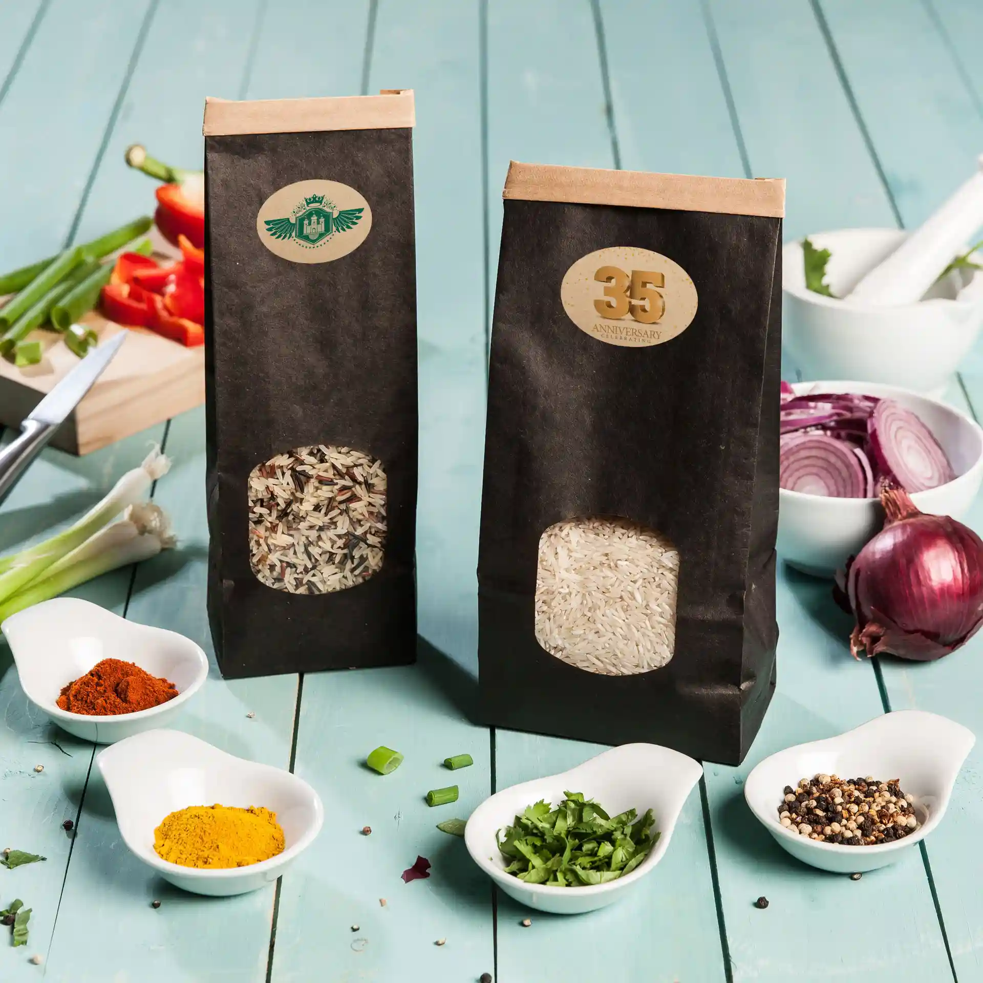 Two bags of rice with transparent windows, surrounded by spices and vegetables on a blue wooden surface.