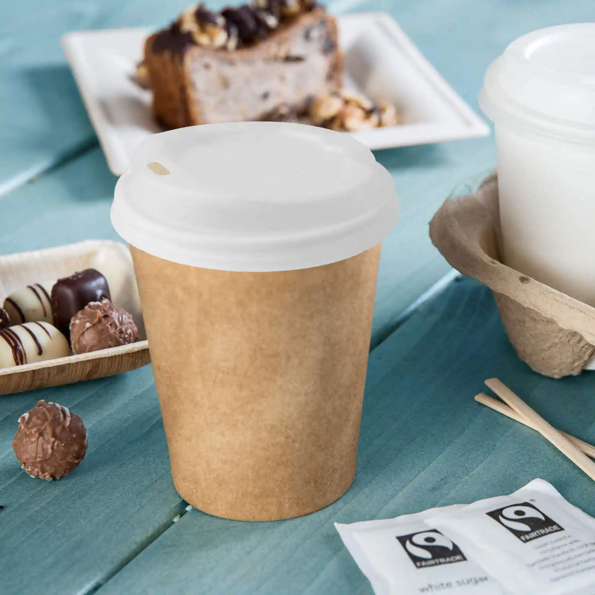 White PS lid with a drinking hole, placed on a brown paper cup, surrounded by desserts and sugar packets.