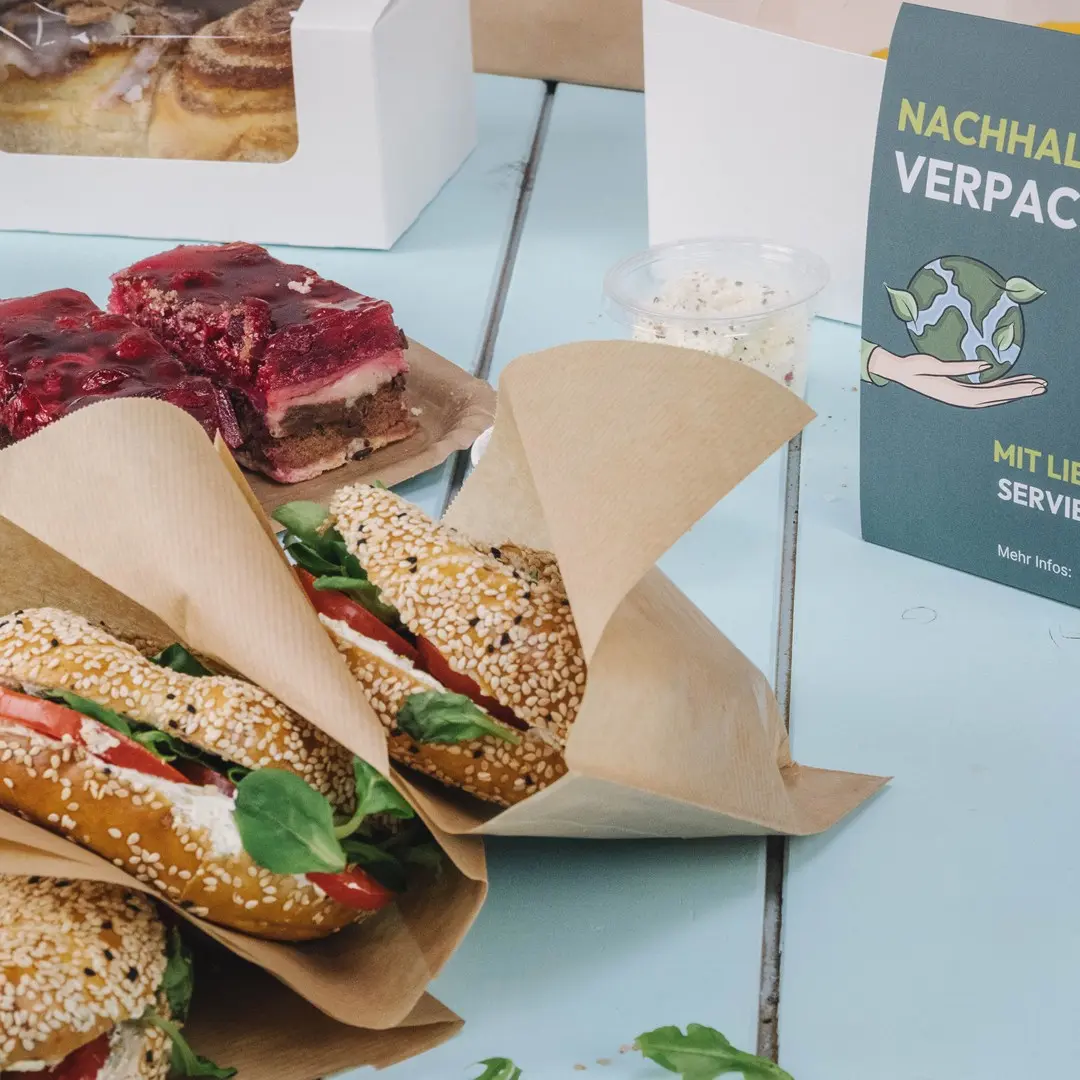 Drei Sesambrötchen gefüllt mit Salat und Tomate, ein Dessert mit Gelee und ein nachhaltigkeitsbezogenes Schild auf einem blauen Tisch.
