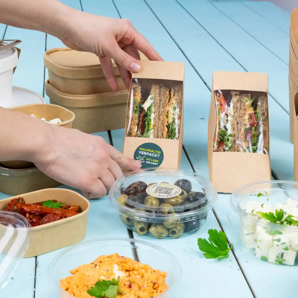 Eine Hand greift nach einem Sandwich-Paket auf einem Tisch; verschiedene Lebensmittelbehälter sind sichtbar, darunter Oliven, ein cremiges Gericht und sonnengereifte Tomaten.