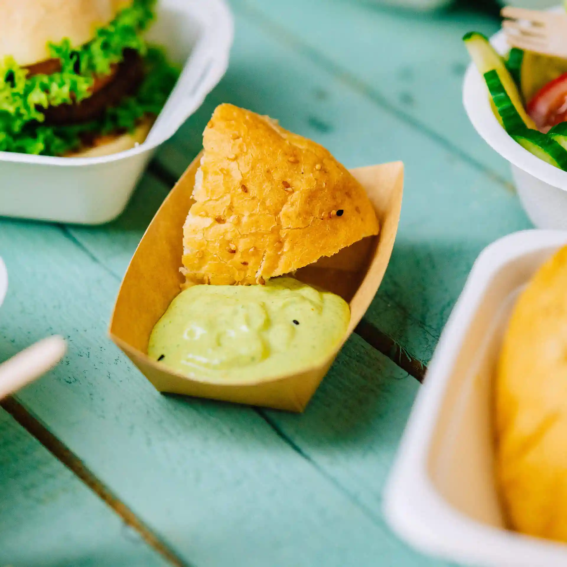 Brown cardboard snack tray containing a piece of fried bread and green sauce on a blue wooden surface.