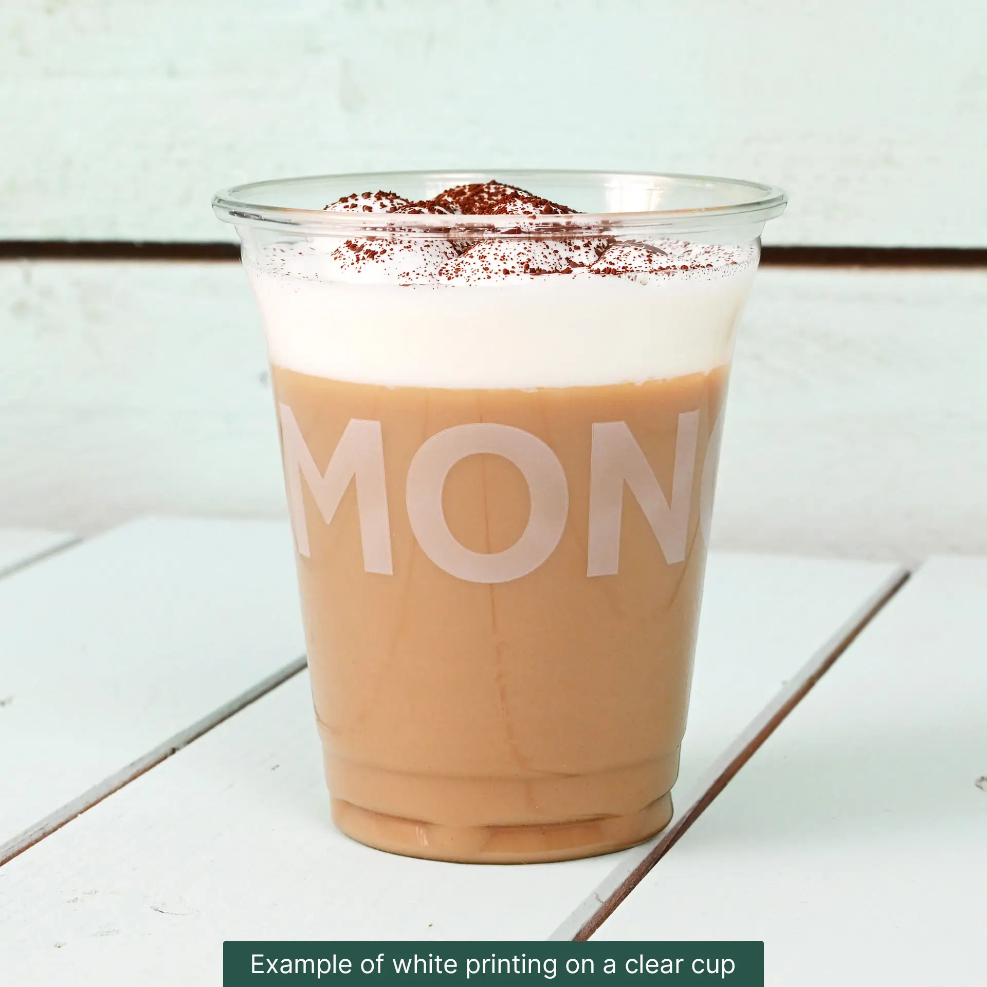 Clear 16 oz rPET cup filled with iced coffee, topped with whipped cream and cocoa powder, placed on a white wooden surface.