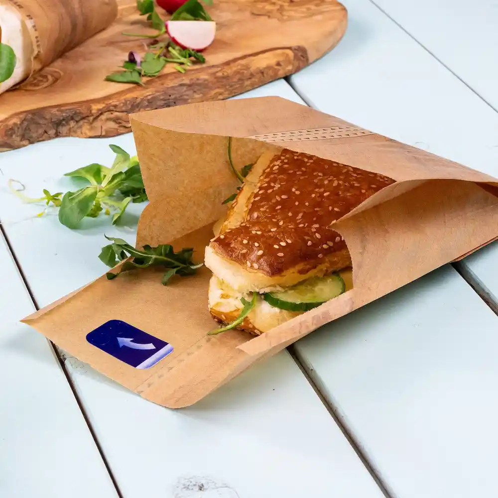 Brown paper bag containing a sesame seed sandwich with cucumbers, surrounded by fresh greens on a blue surface.