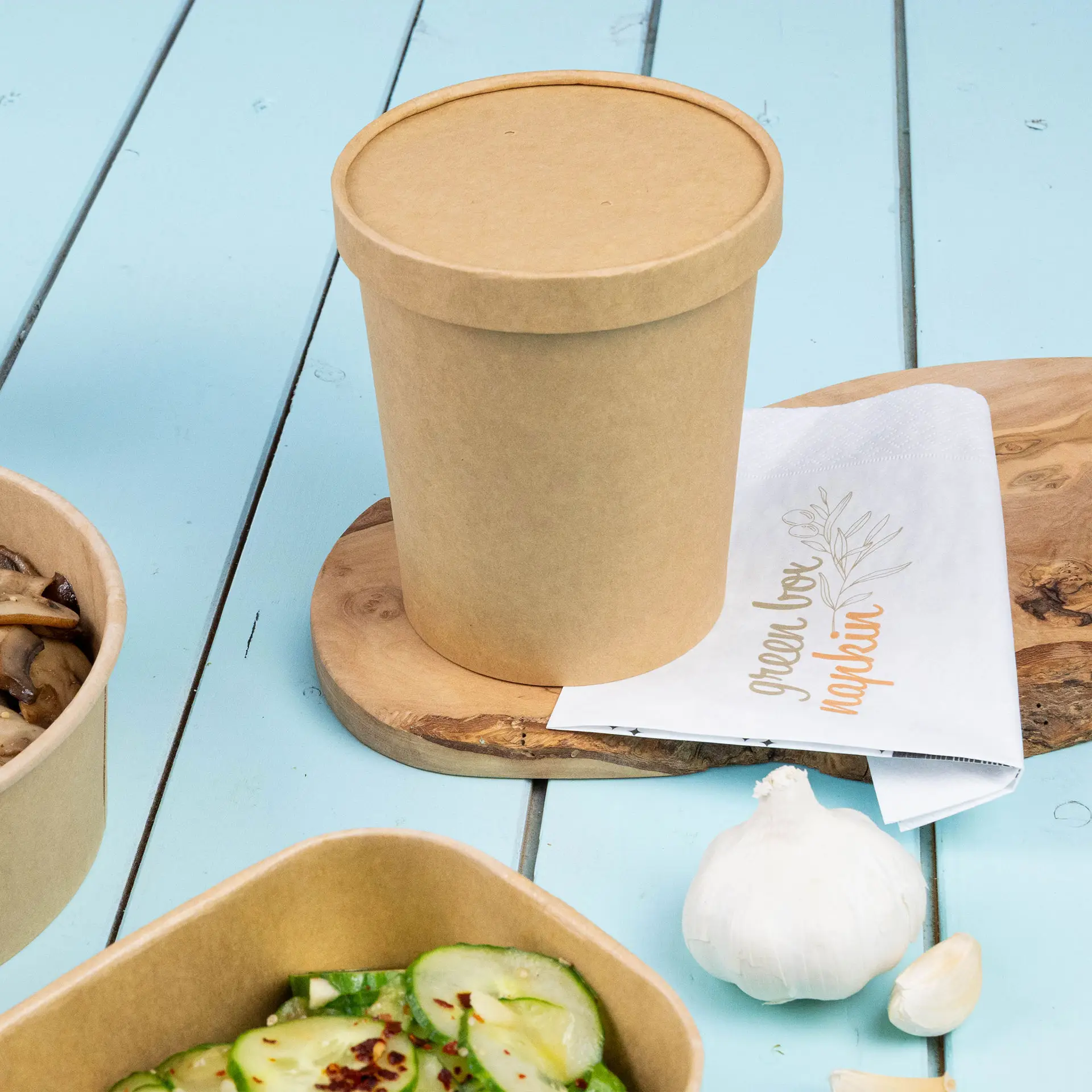 Brauner Pappbecher für Suppe mit Deckel, auf einem Holzbrett, neben einer Serviette und Lebensmittelbehältern.