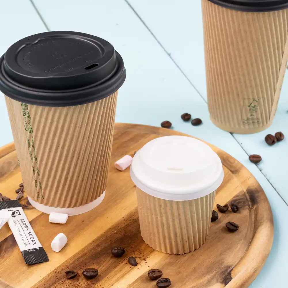 Drei Papierbecher in verschiedenen Größen mit Deckeln, auf einem Holztablett mit Kaffeebohnen und Zuckertütchen.
