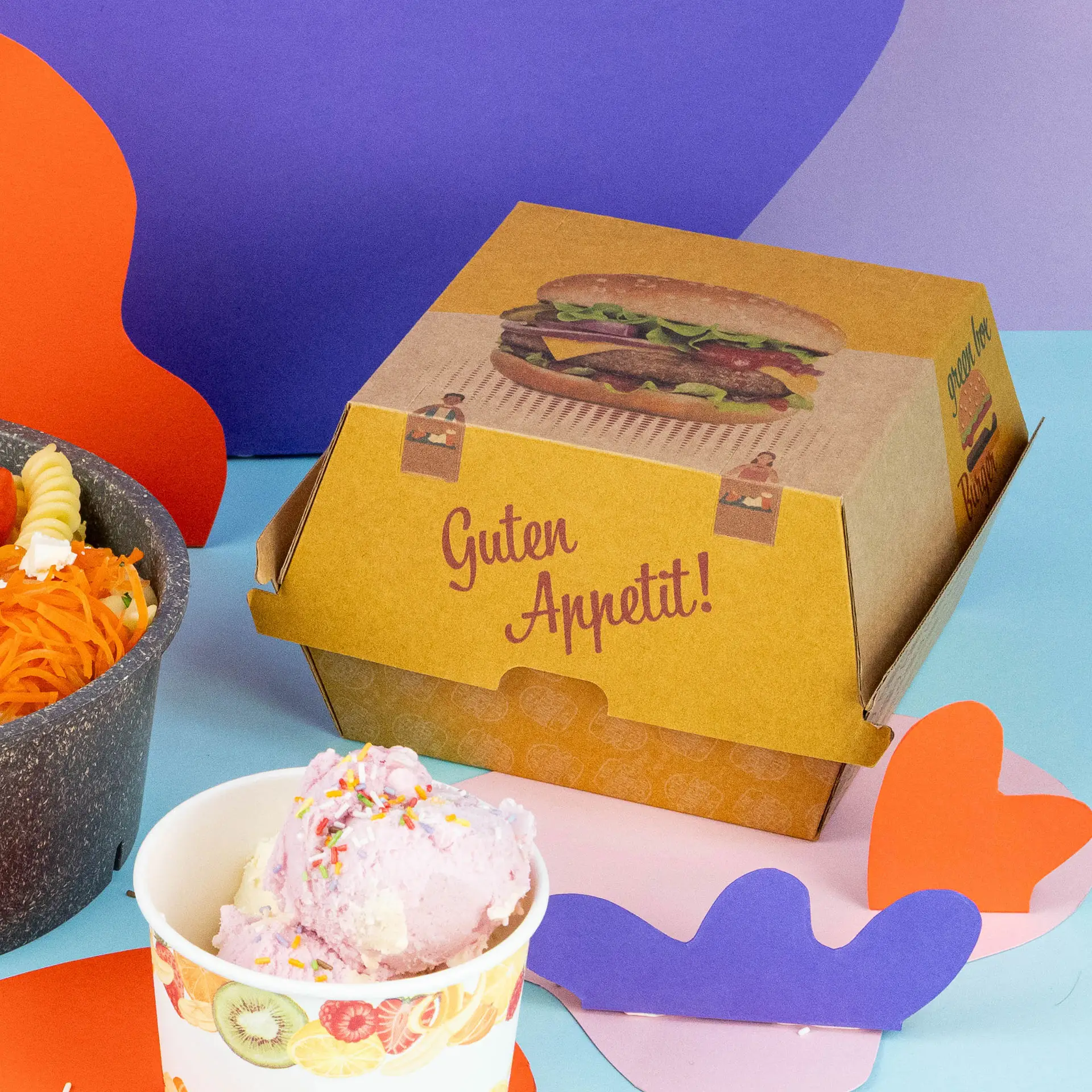 One whole burger with sesame seeds in a kraft cardboard box, alongside a bowl of mixed vegetables and a small sauce container.