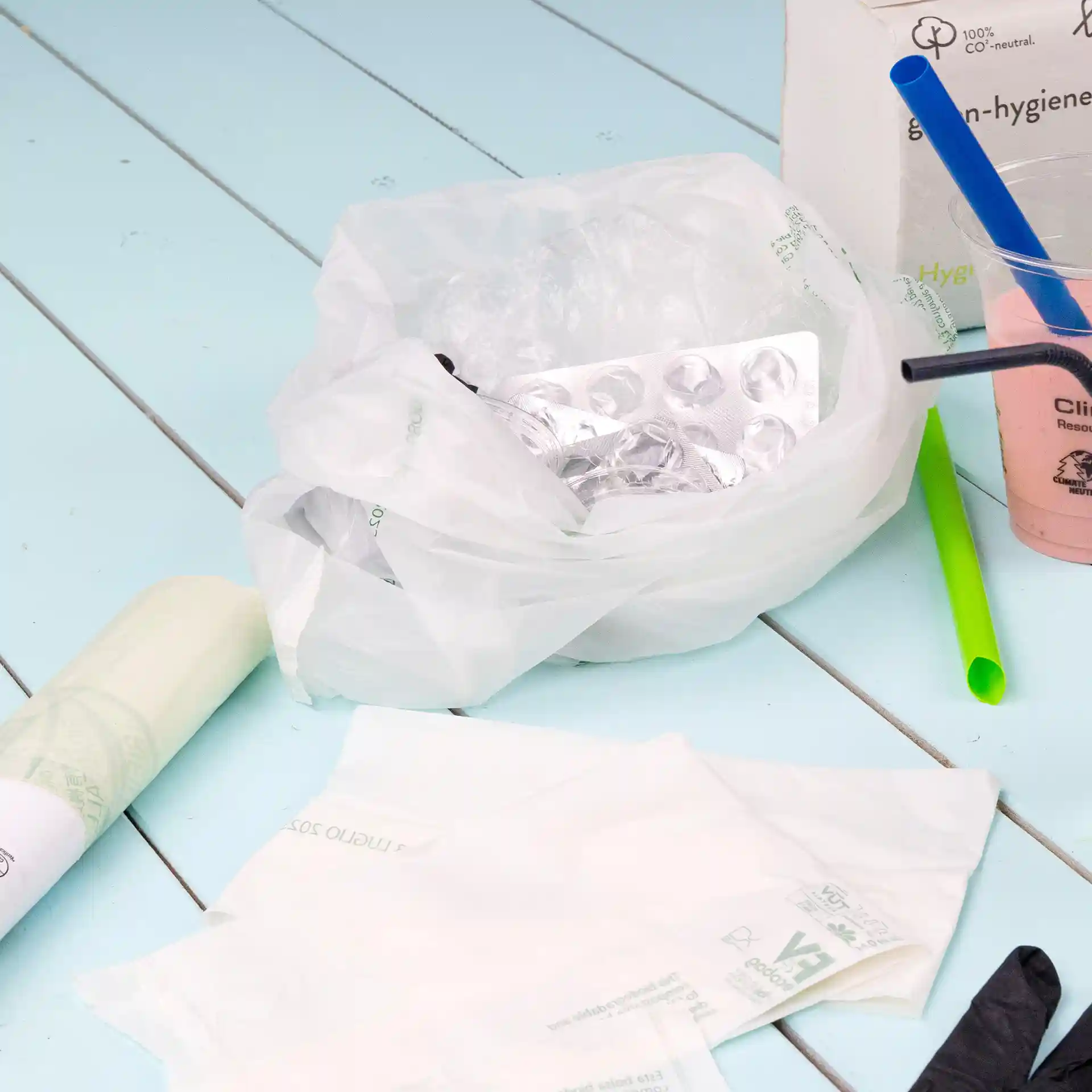 Transparente Bio-Mülltüte mit Inhalt, darunter eine Blisterverpackung, Plastikstrohhalme und Einwegartikel auf blauer Oberfläche.