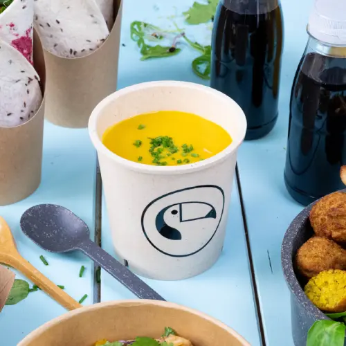 Suppenbecher mit gelber Suppe und grünem Zubehör, umgeben von Lebensmittelbehältern und Getränken auf einer blauen Oberfläche.