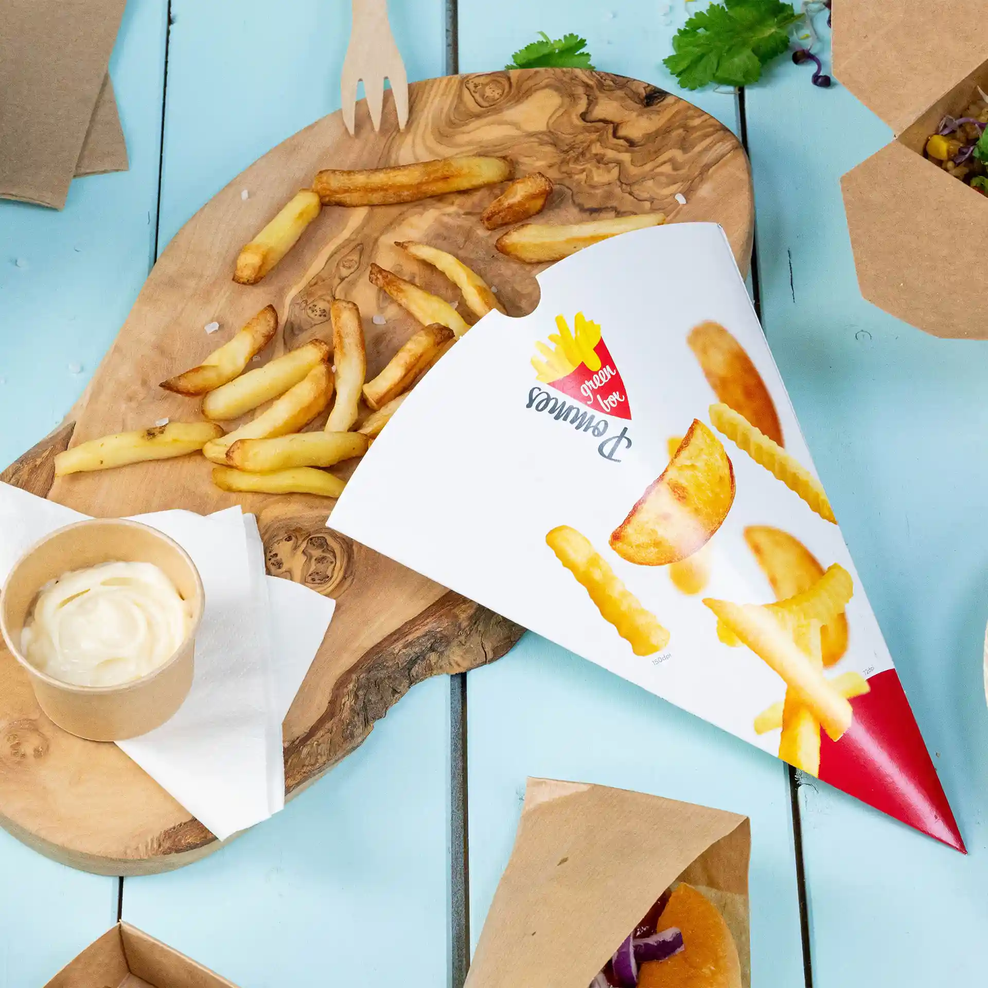 Printed french fry cone with fries, a cup of sauce, and a wooden serving board on a blue surface.