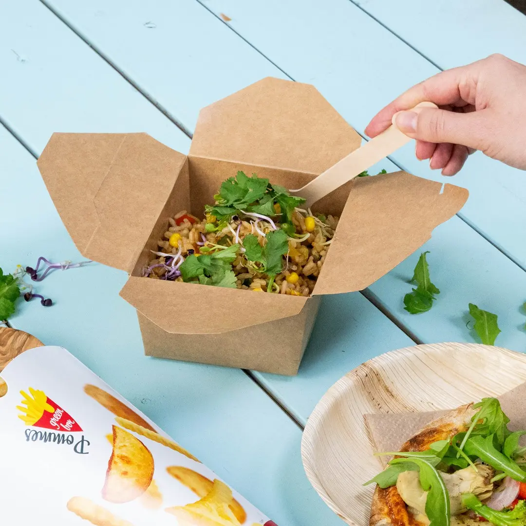 Brown takeout box filled with mixed vegetables and herbs, with a hand holding a wooden spoon, on a blue table.