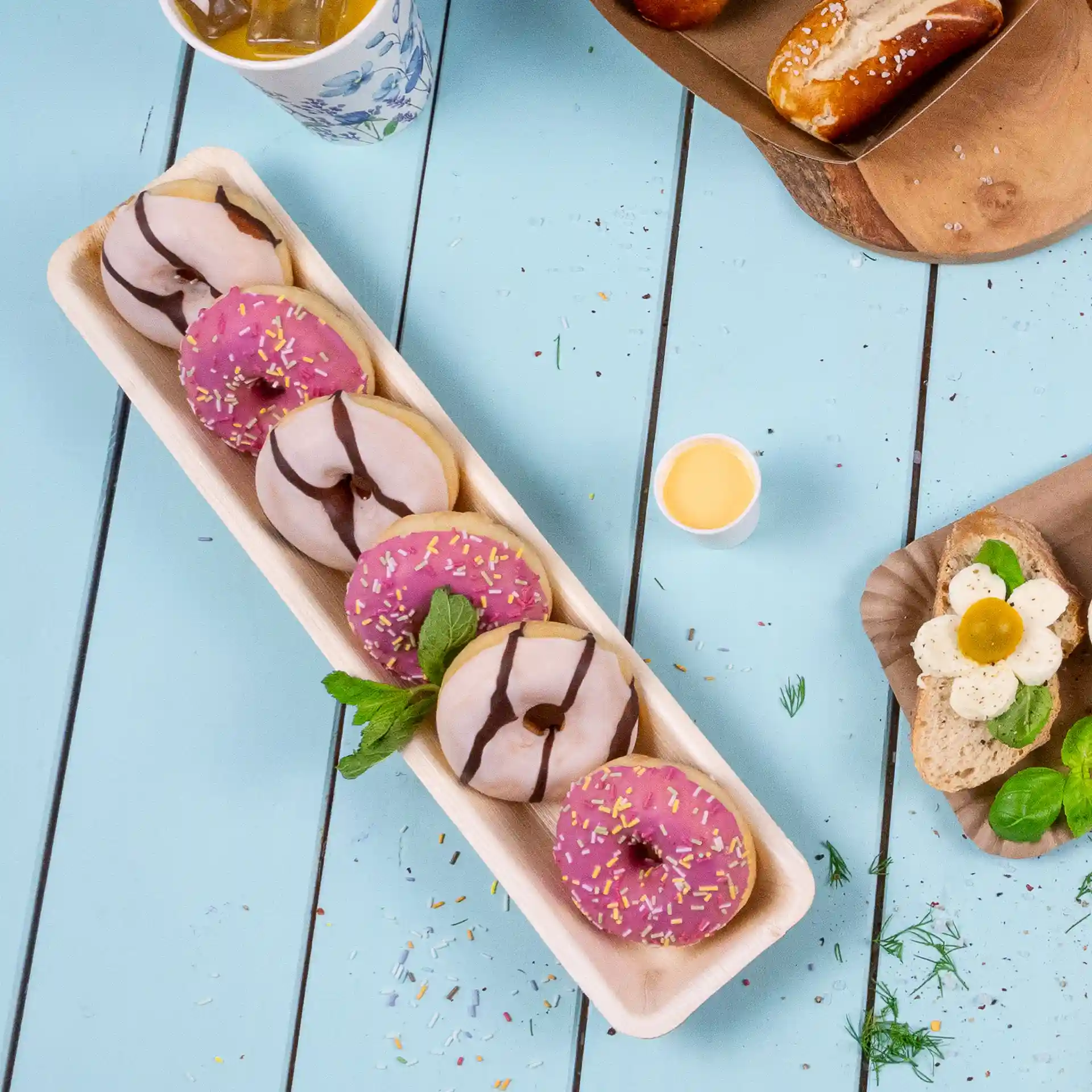 Rectangular palm leaf tray with several donuts, including pink and chocolate varieties, on a blue wooden surface.