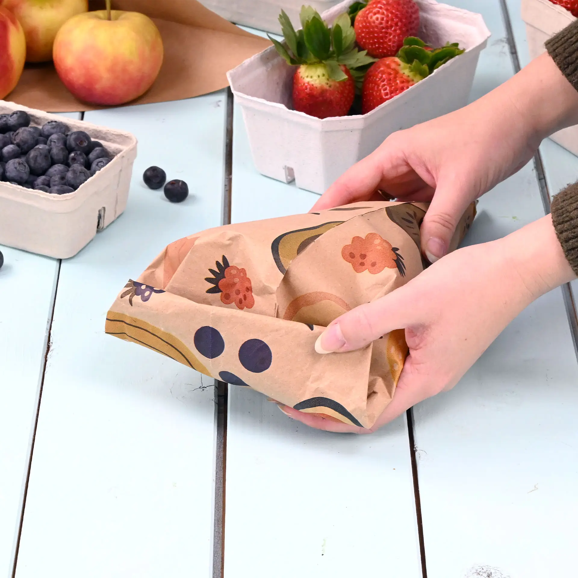 Hands holding a custom printed paper cone with a brown design, surrounded by fruit baskets on a wooden table.