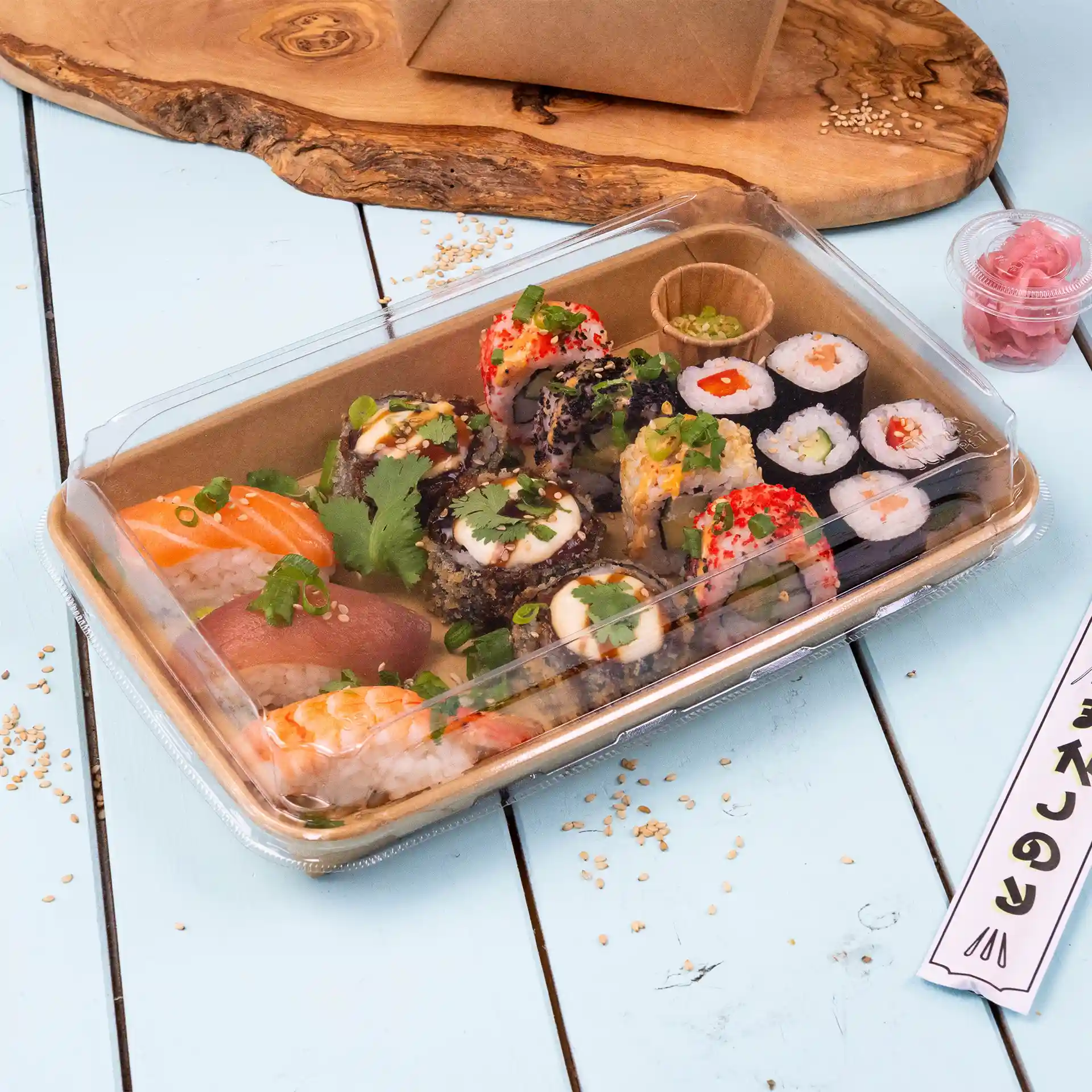 Transparenter Sushi-Box-Deckel mit verschiedenen Sushi-Rollen, darunter Lachs, Thunfisch und Beilagen, auf einer hellblauen Oberfläche.